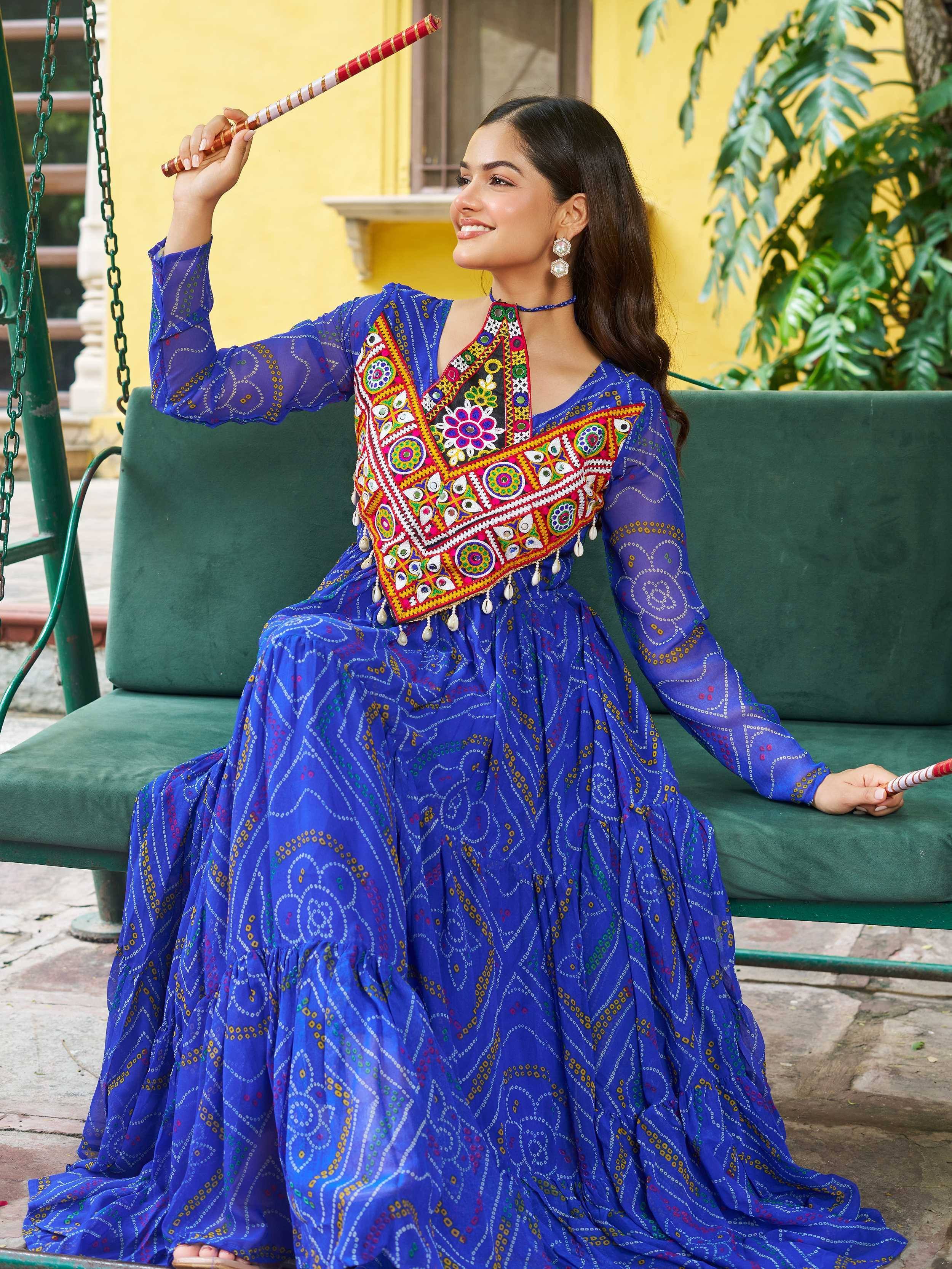 Woman in vibrant blue embroidered ethnic gown with mirror work, holding dandiya stick, seated on green swing