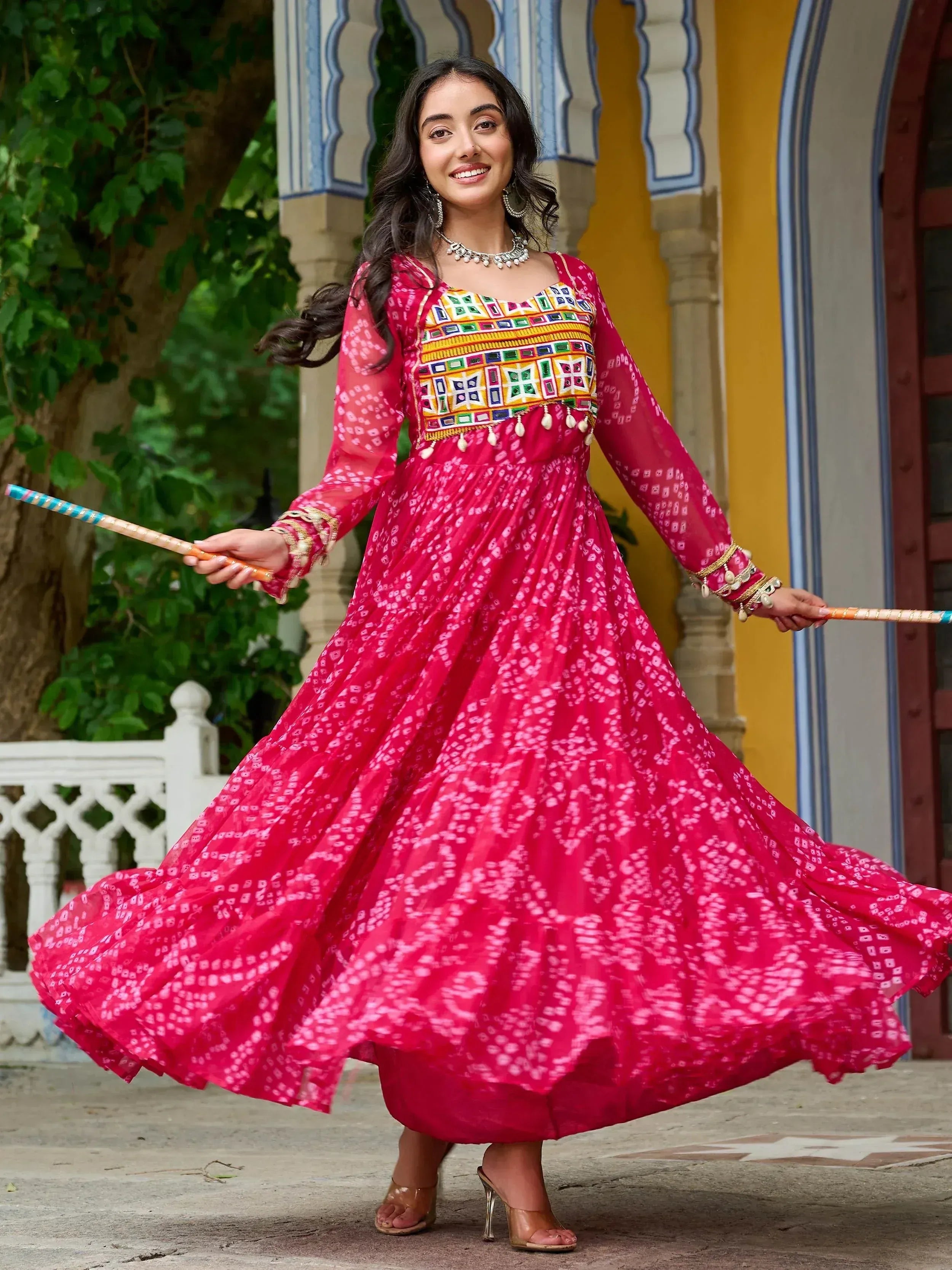 Woman in vibrant pink ethnic gown with mirror work, twirling with dandiya sticks, perfect for festive occasions.