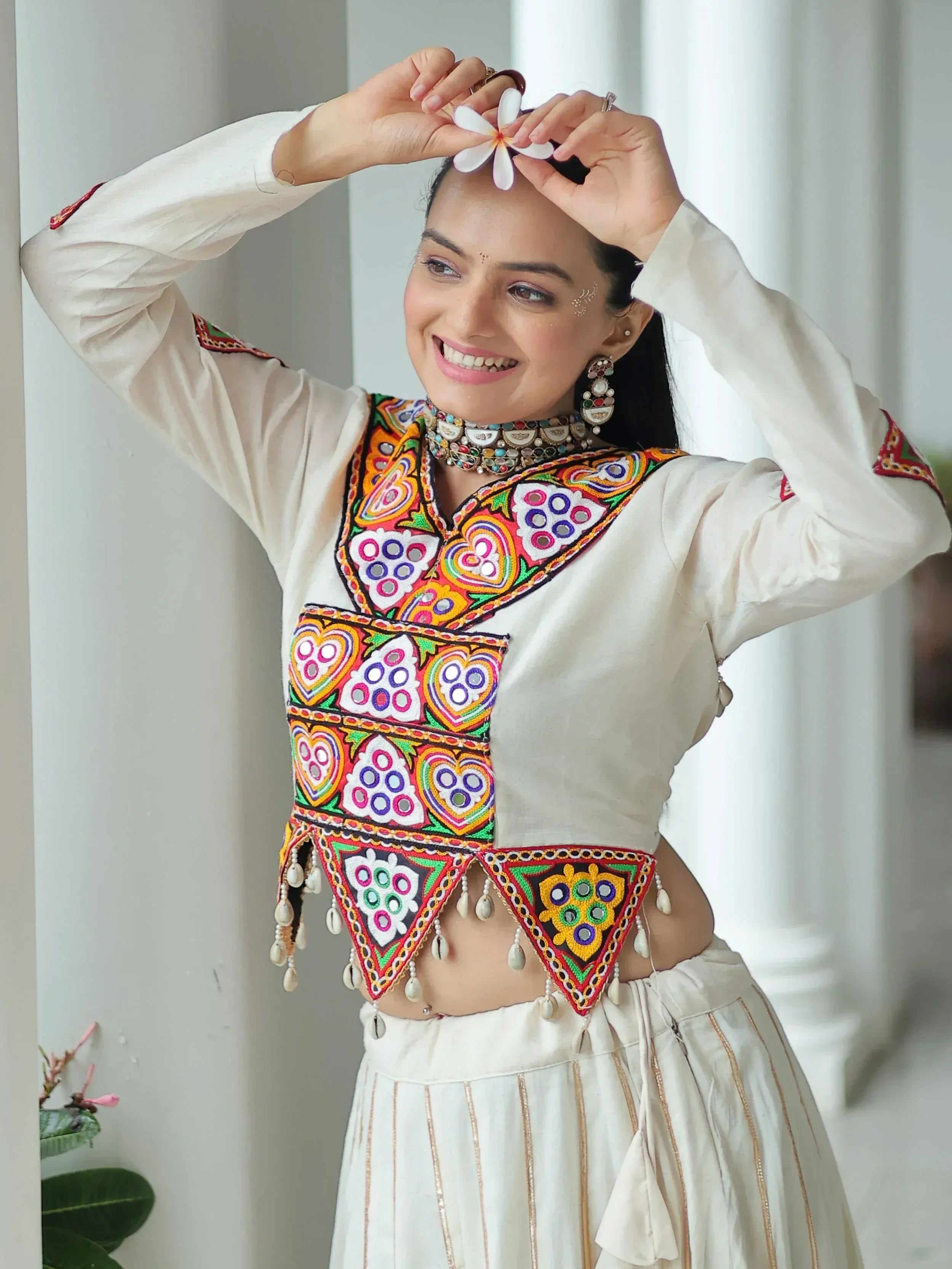 Woman in white embroidered ethnic top with mirror work, floral jewelry, and gold-trimmed skirt, holding white flower.