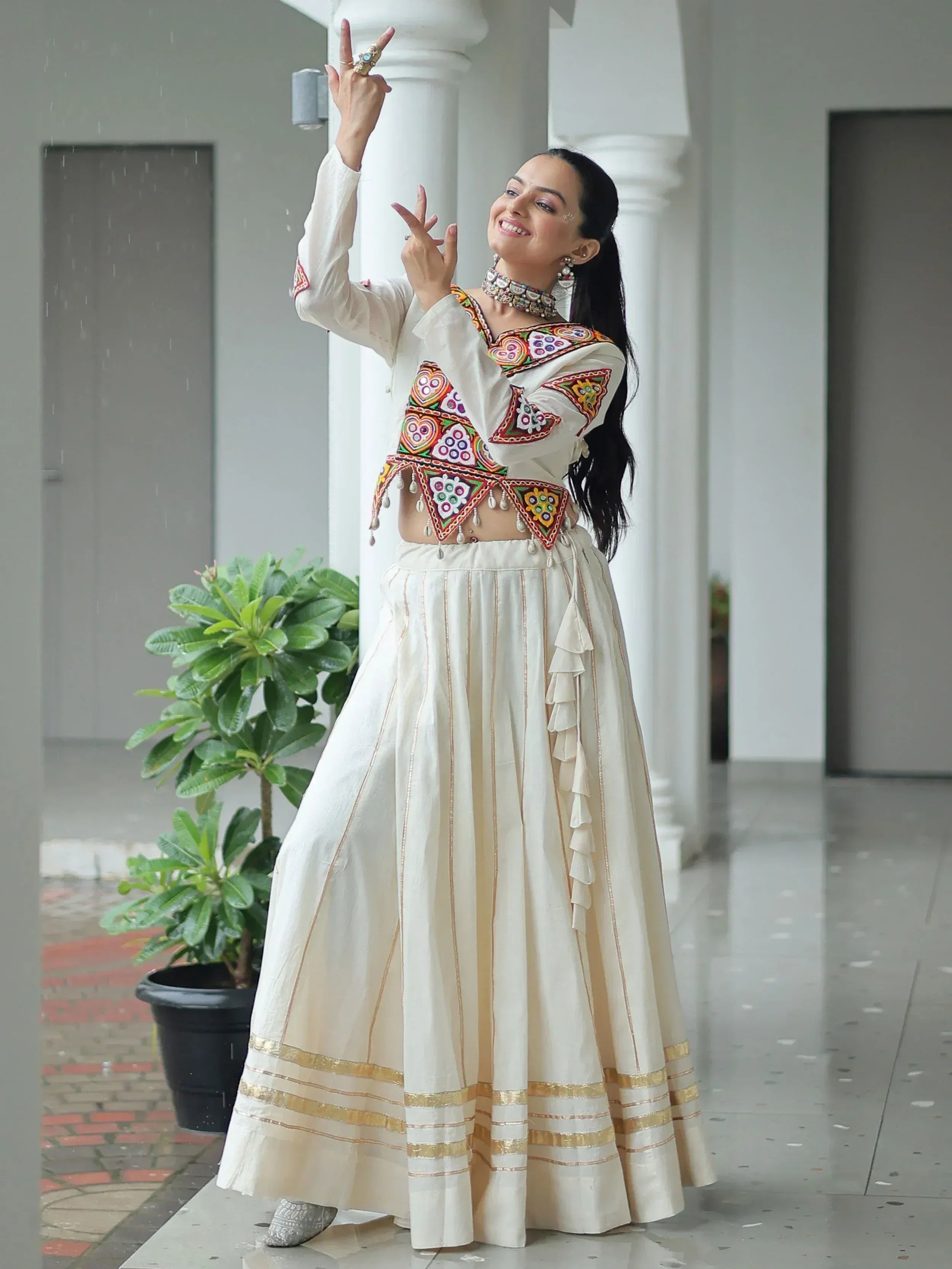 Woman in white embroidered lehenga choli with colorful mirror work, dancing joyfully in rain under white pillars