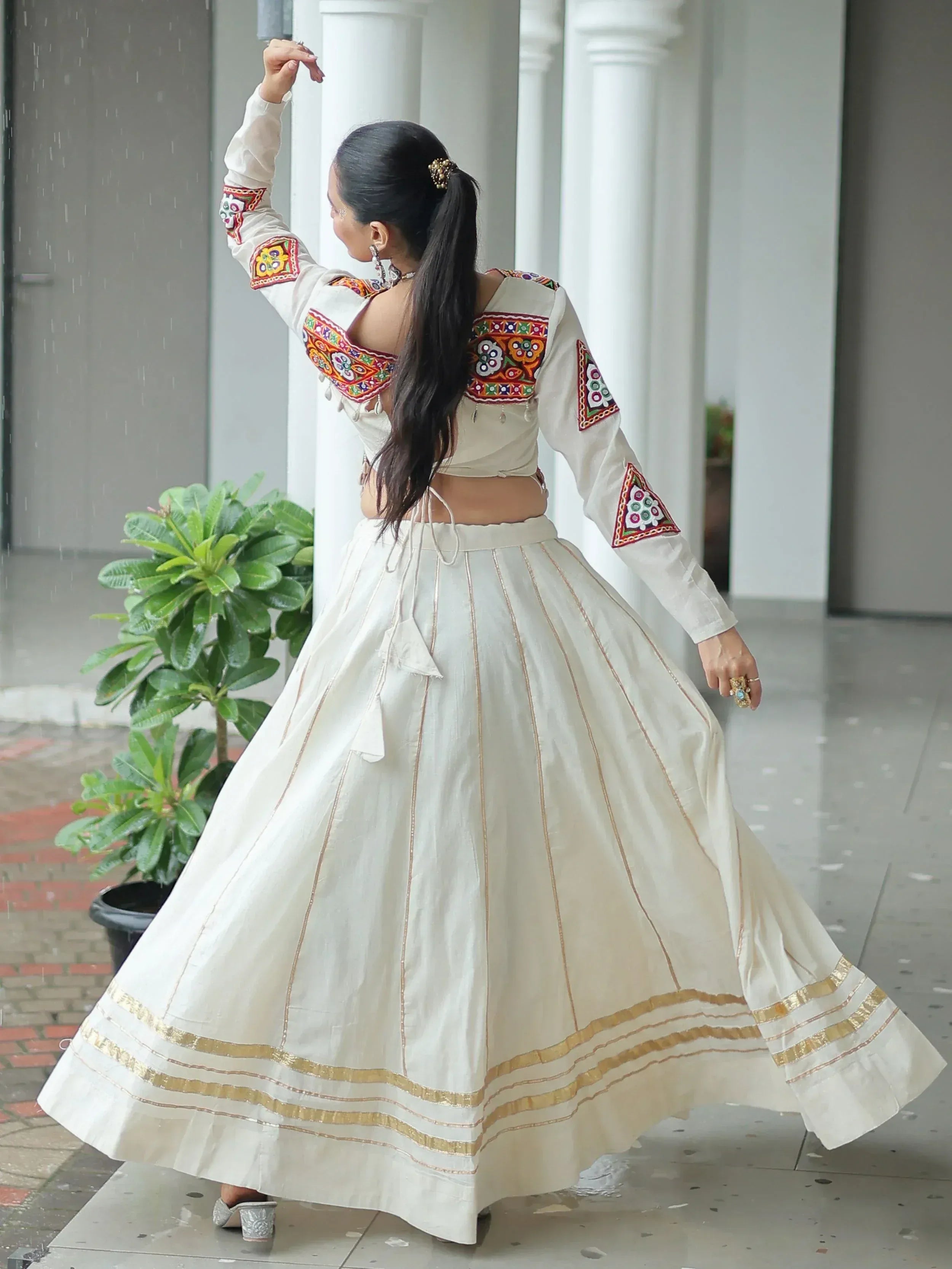 Woman in white embroidered lehenga choli with gold border, dancing in rain under white pillars