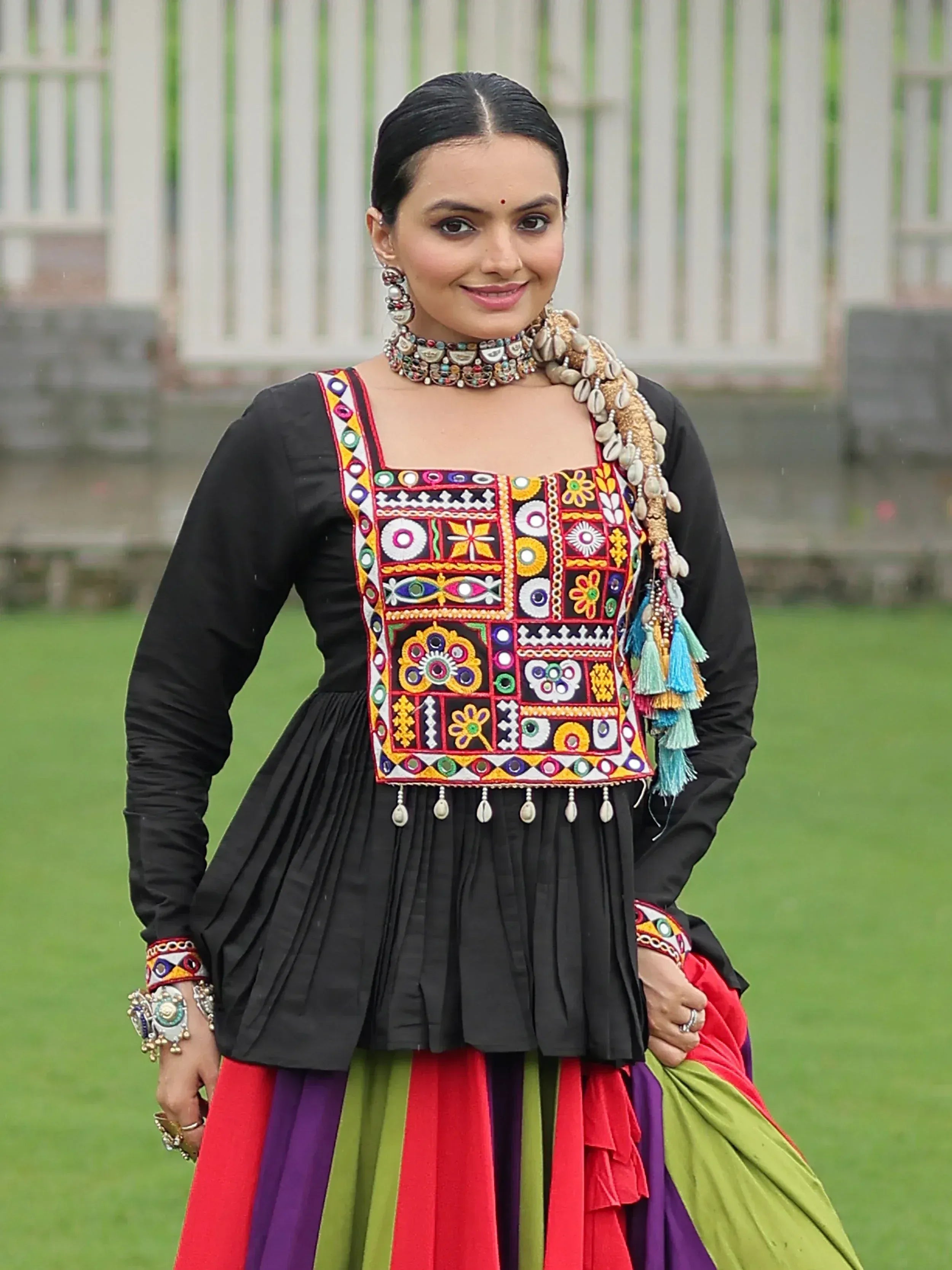 Woman in black embroidered ethnic top with colorful mirror work, paired with multicolor skirt and traditional jewelry.