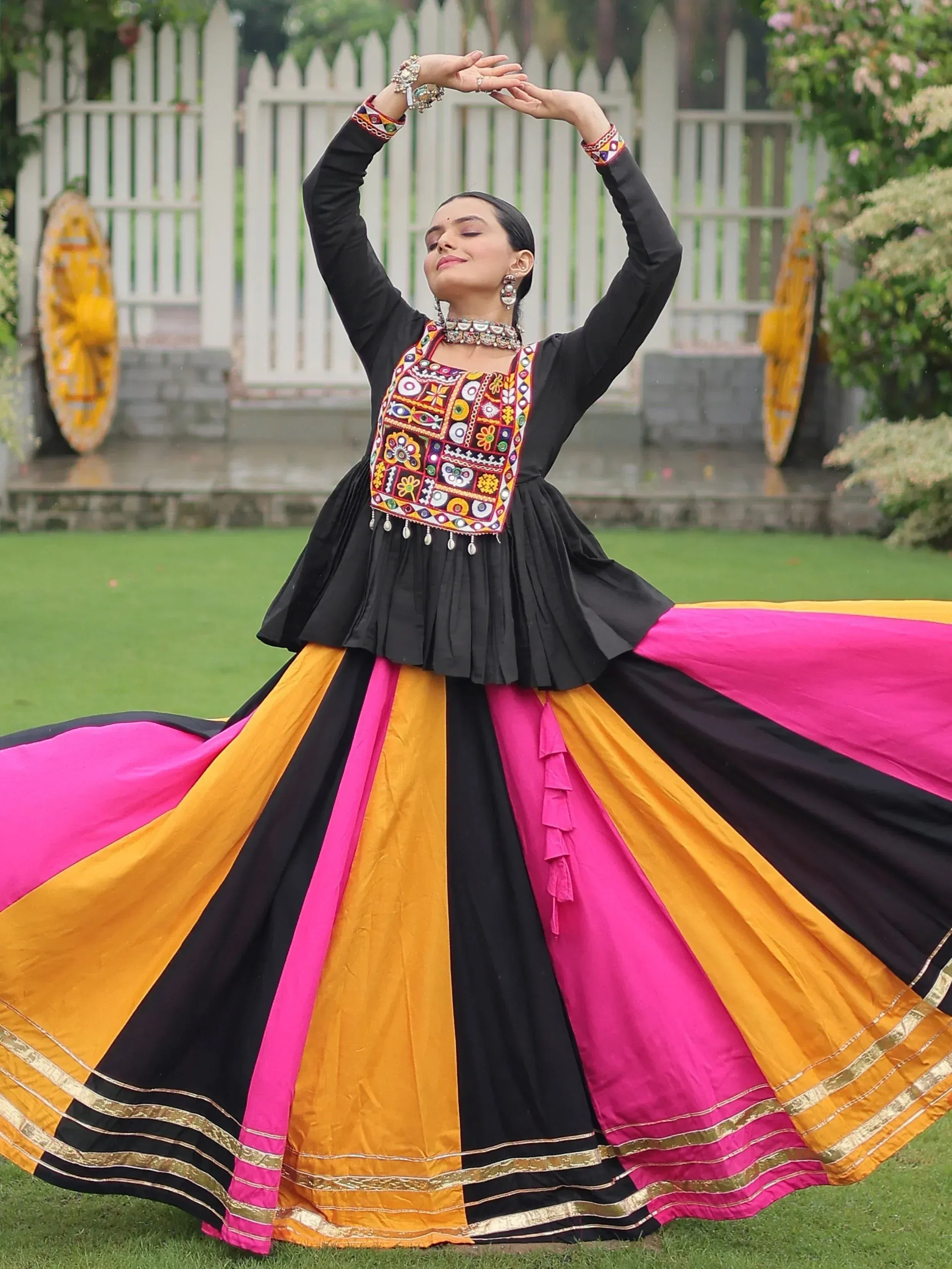 Woman in vibrant black, pink, and yellow ethnic gown with mirror work, twirling outdoors