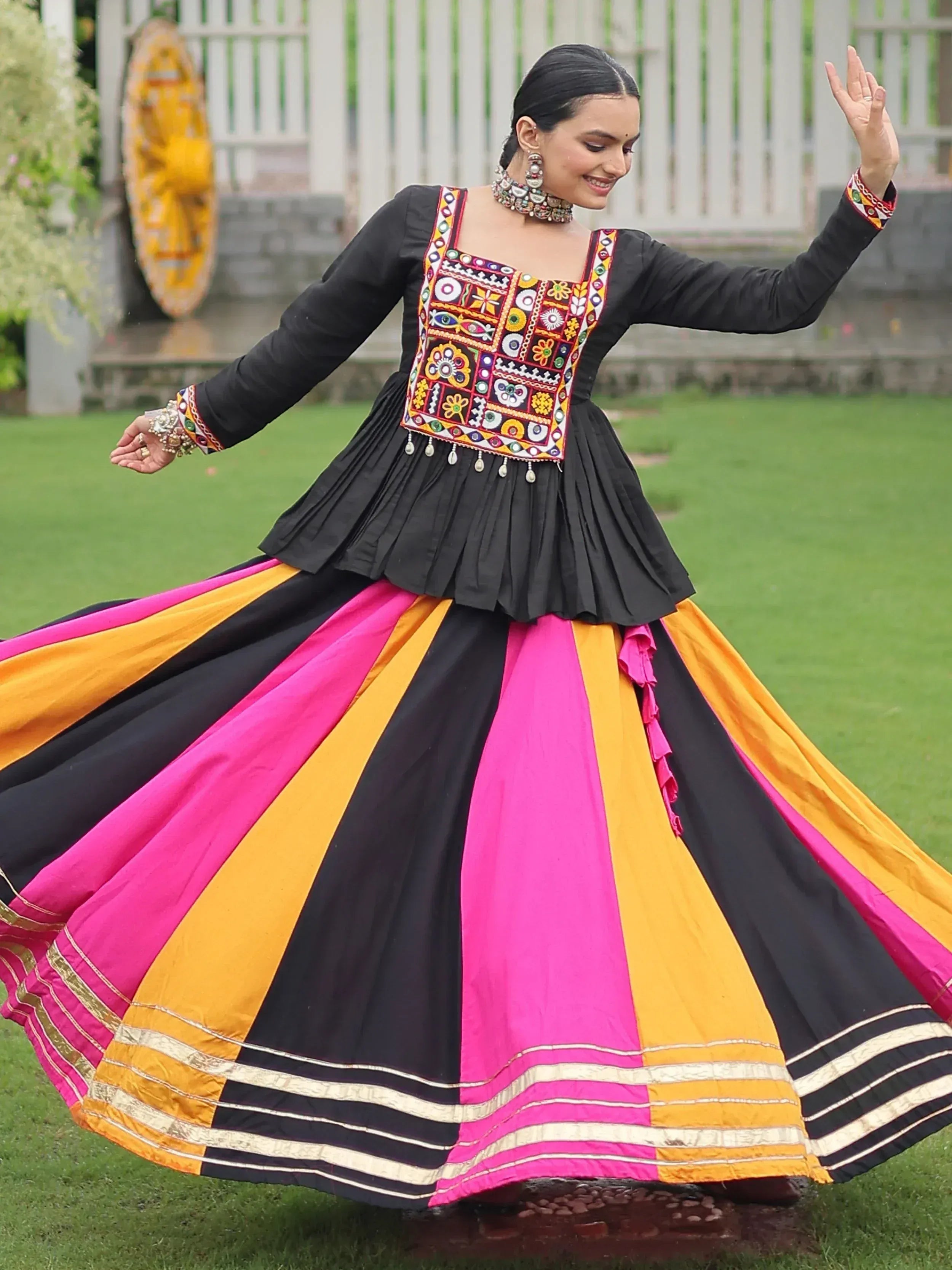 Woman in black, pink, and yellow ethnic gown with mirror work, twirling outdoors