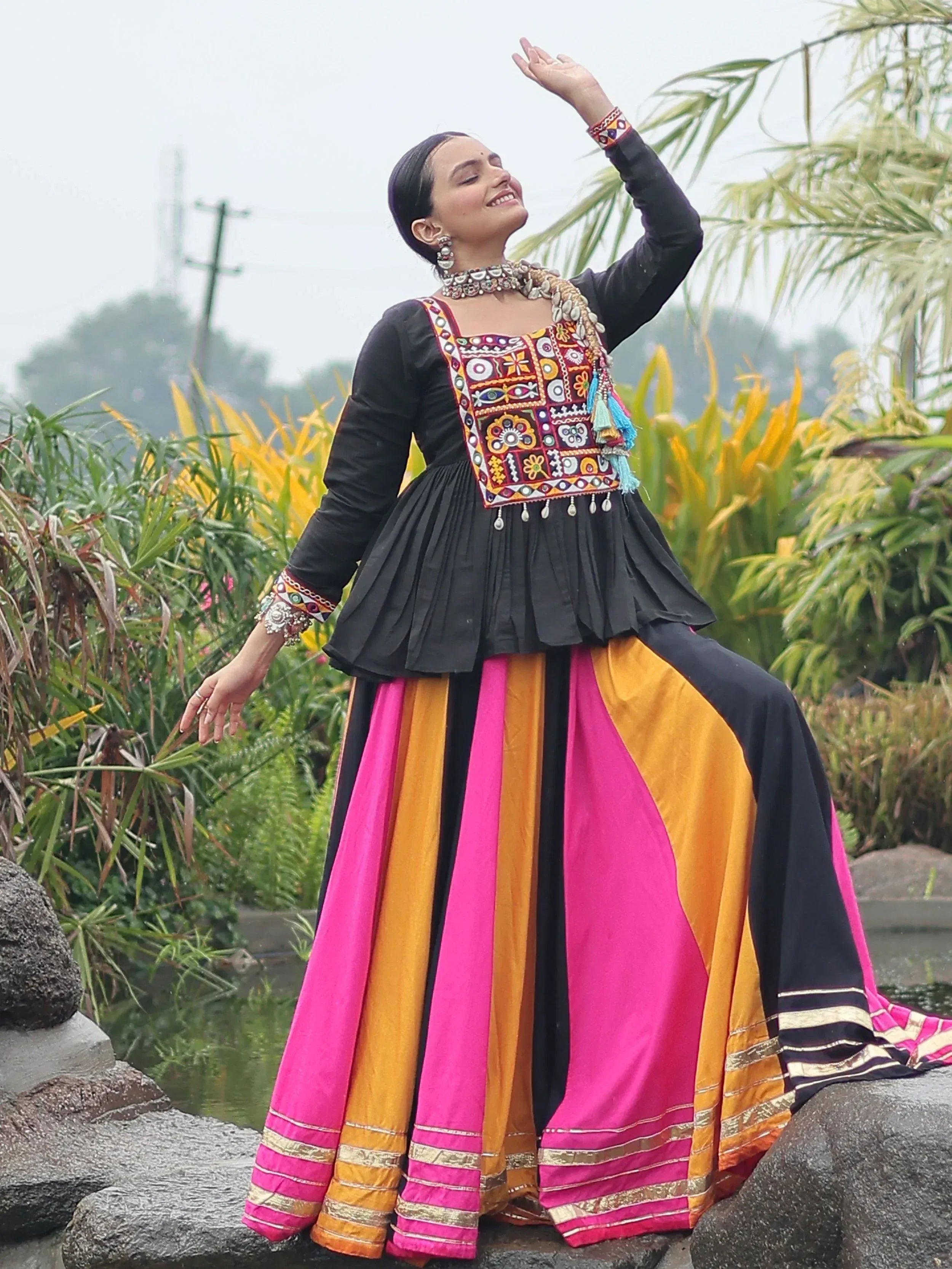 Woman in vibrant ethnic gown with embroidered black top and pink-orange skirt, posing outdoors