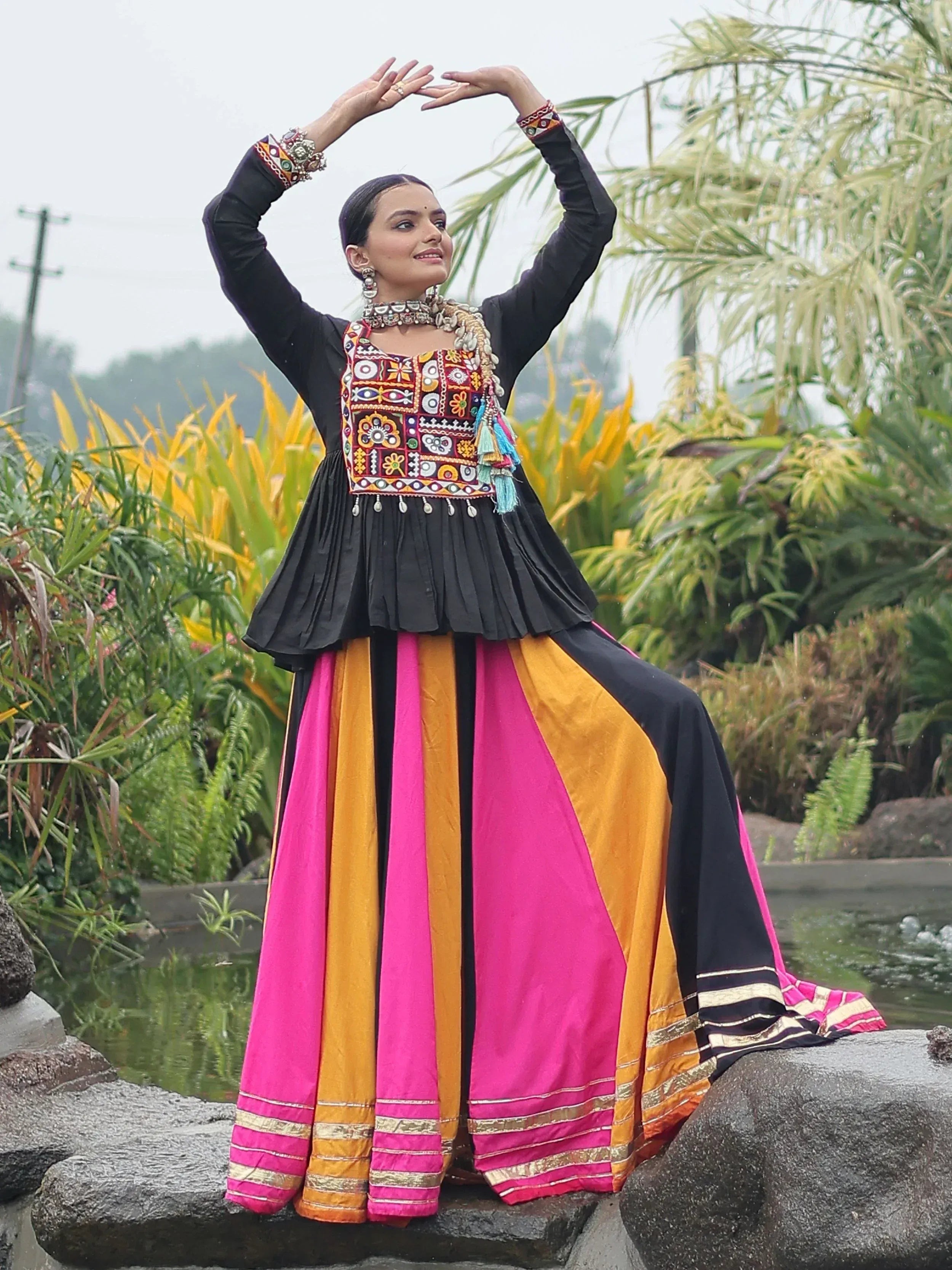 Woman in vibrant ethnic gown with colorful embroidery and pleated skirt, posing outdoors
