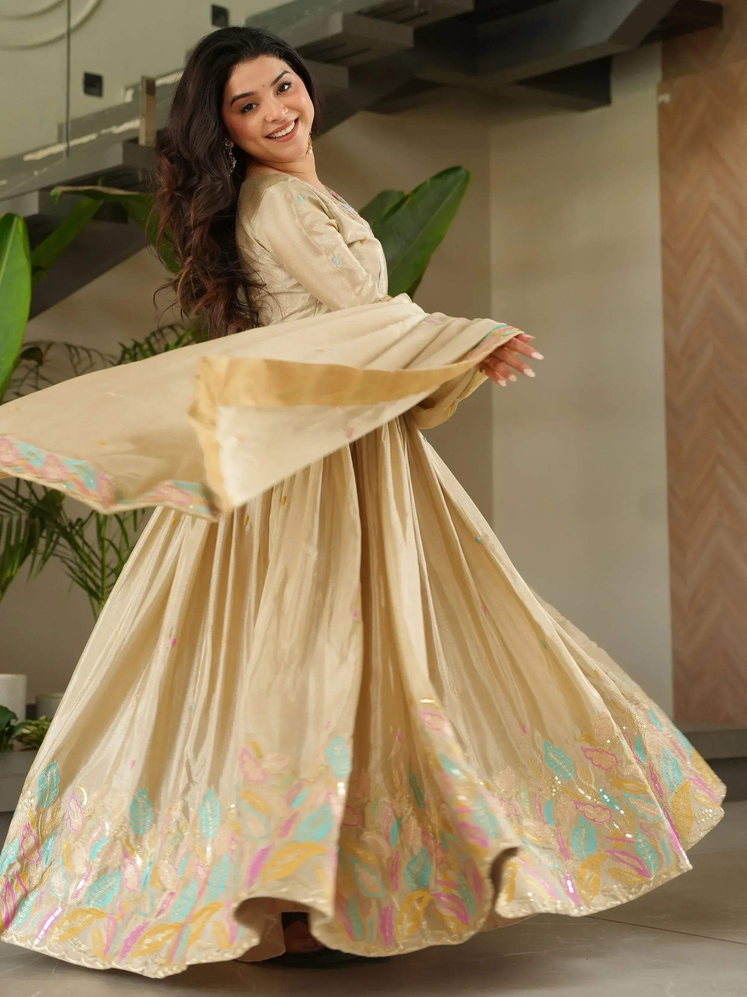 Woman twirling in elegant beige ethnic gown with colorful floral embroidery, perfect for Indian celebrations.