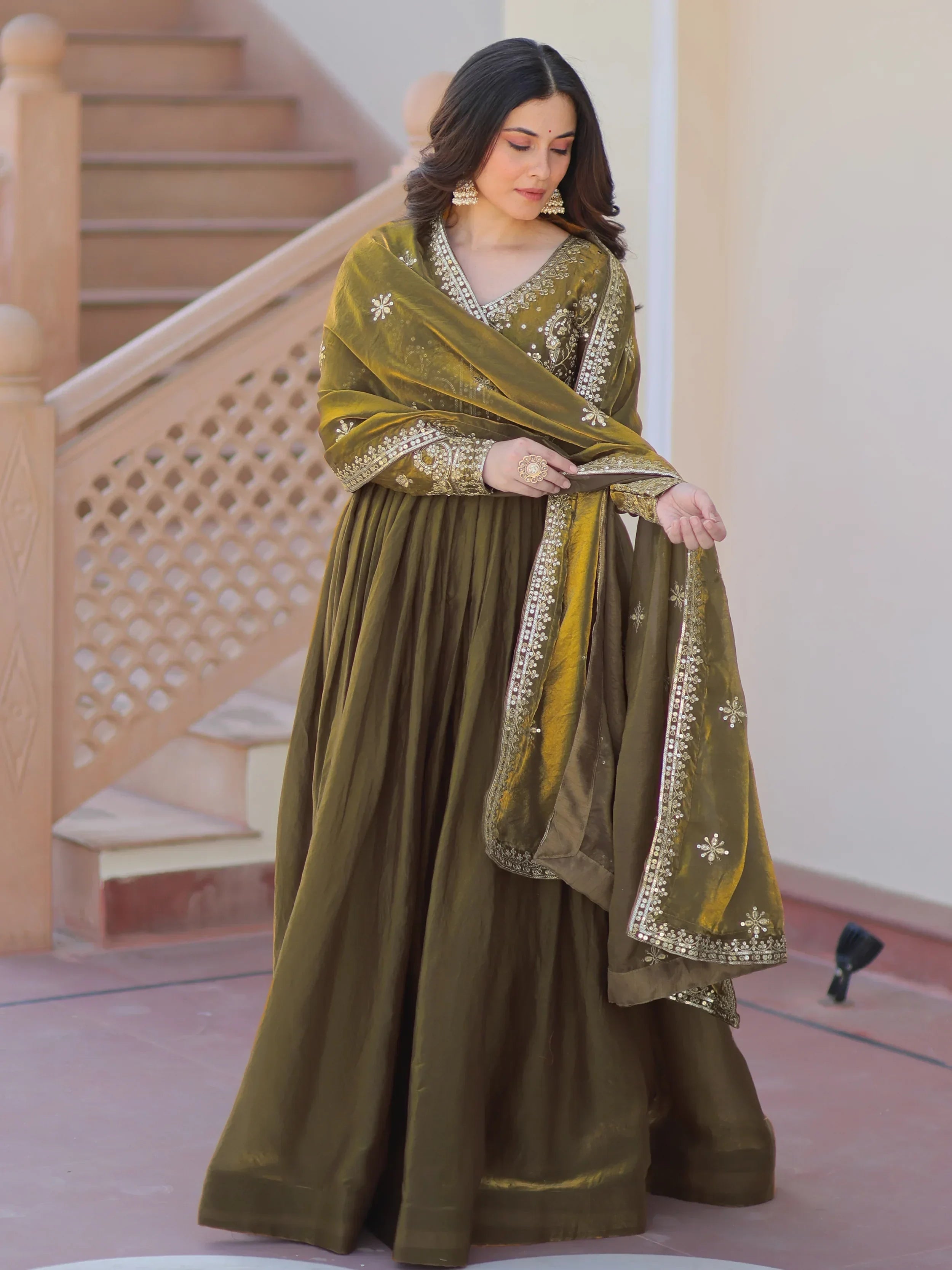 Woman in olive green embroidered ethnic gown with matching dupatta, standing by stairs
