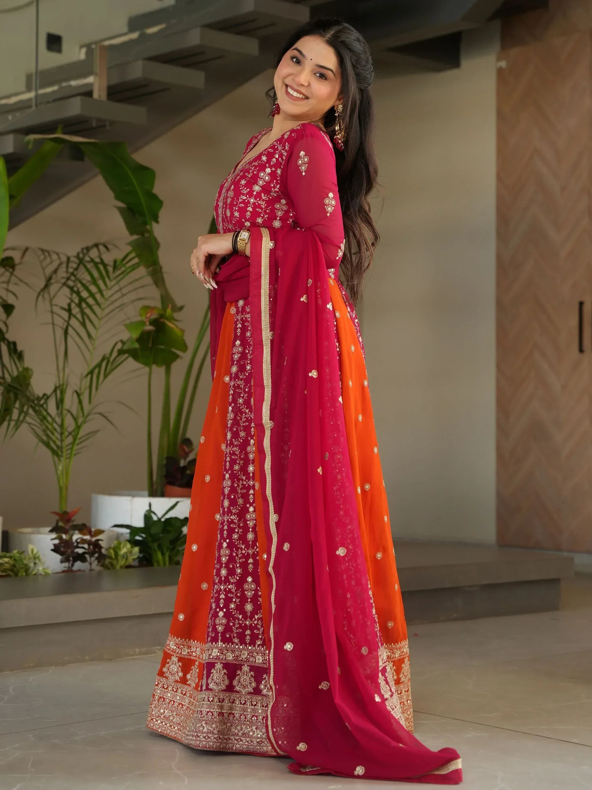 Woman in vibrant pink and orange embroidered Indian ethnic gown with gold detailing, perfect for festive occasions.