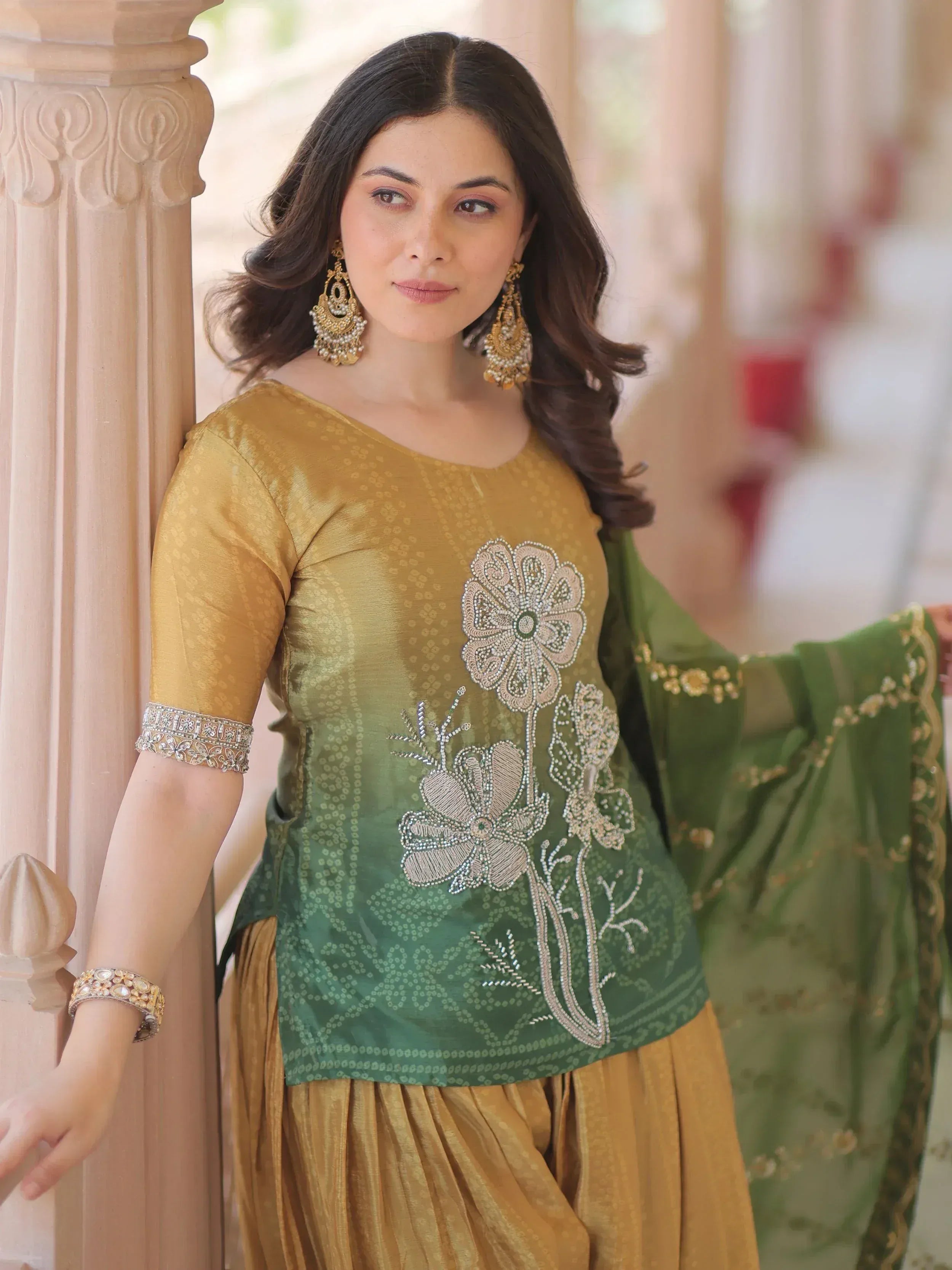 Woman in gold and green ethnic salwar kameez with floral embroidery and dupatta, styled with jhumka earrings.
