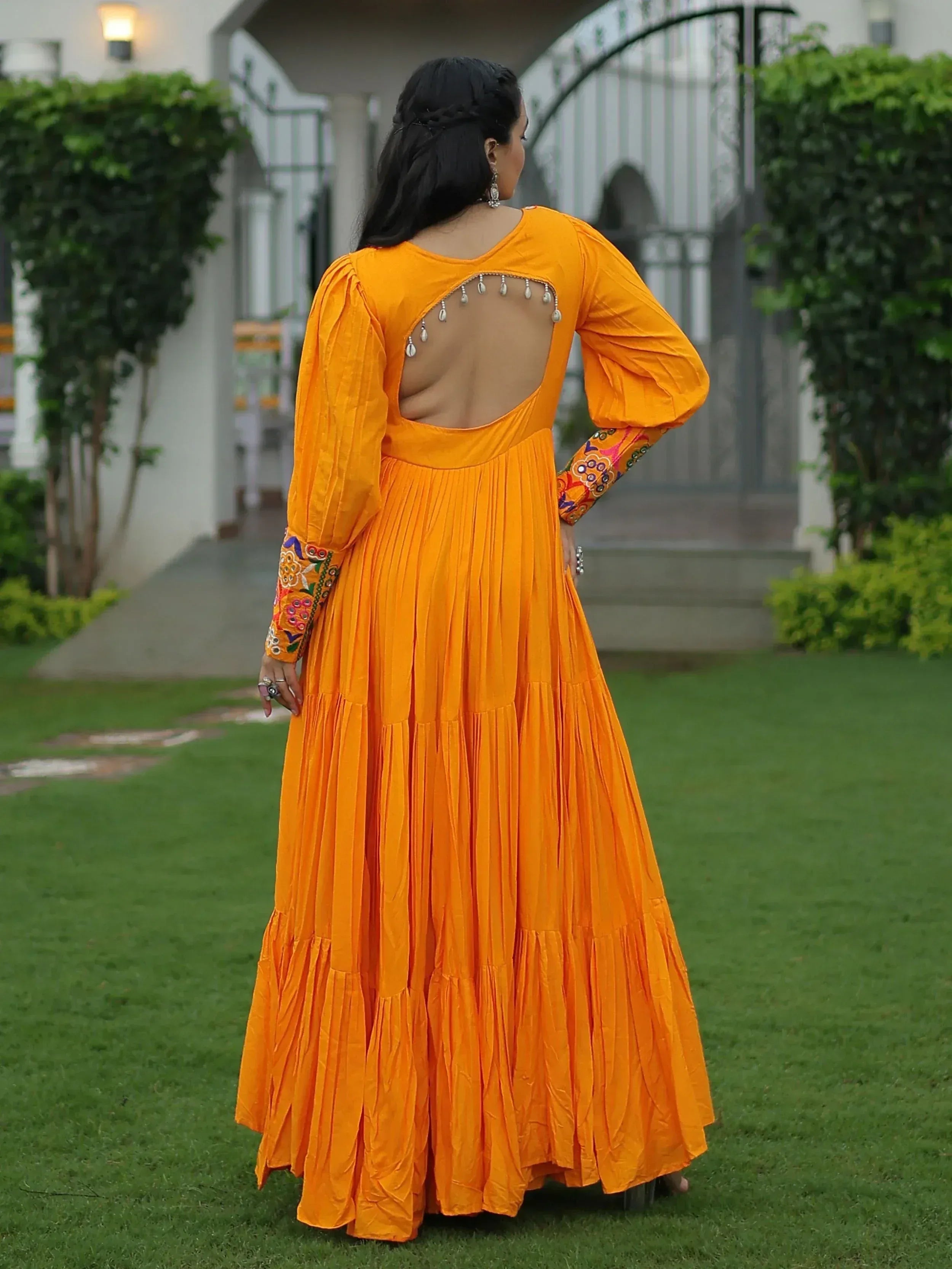 Vibrant orange ethnic gown with back cutout, mirror work, and embroidered sleeves, perfect for Indian festive occasions.