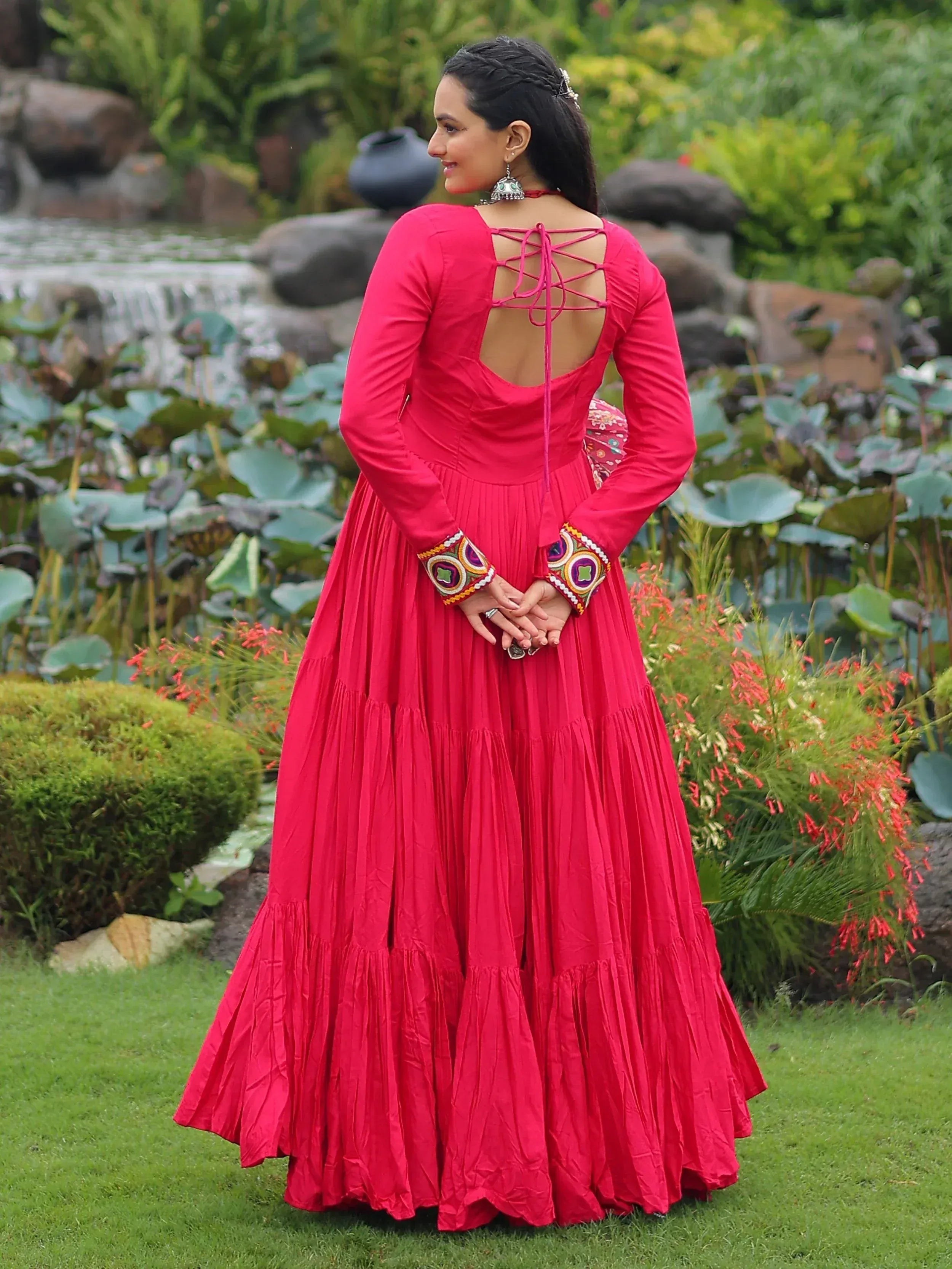 Woman in vibrant red ethnic gown with lace-up back and embroidered sleeves, standing in garden