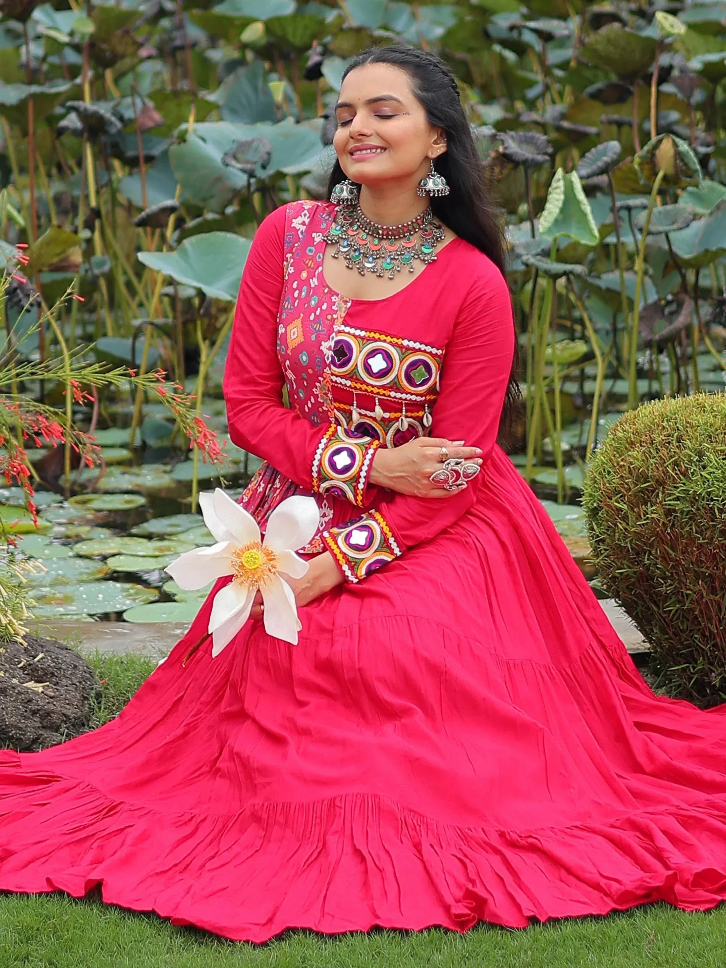 Woman in vibrant pink embroidered ethnic gown with mirror work, holding white lotus, garden setting