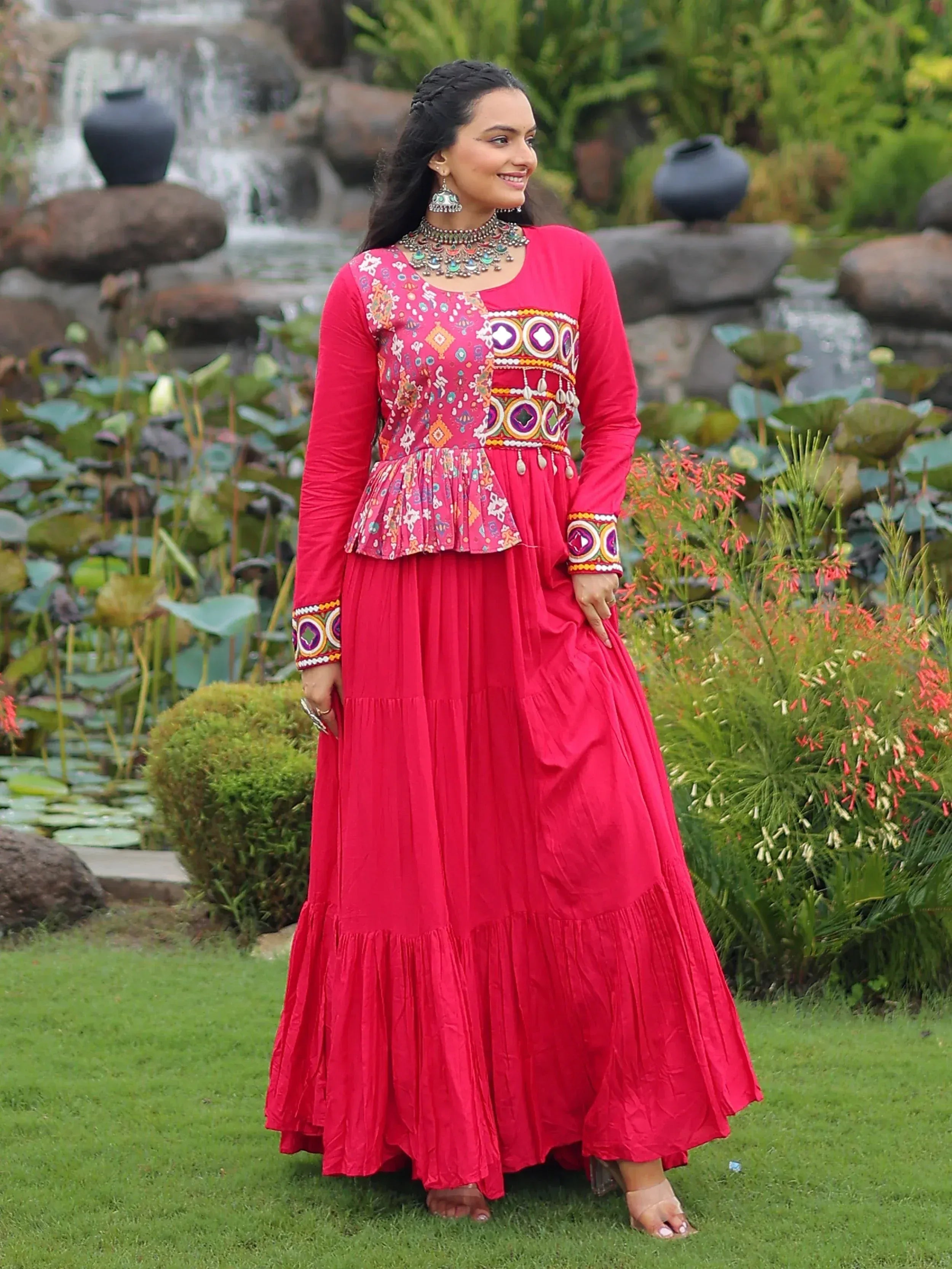 Woman in vibrant red ethnic gown with mirror work, standing in garden near waterfall