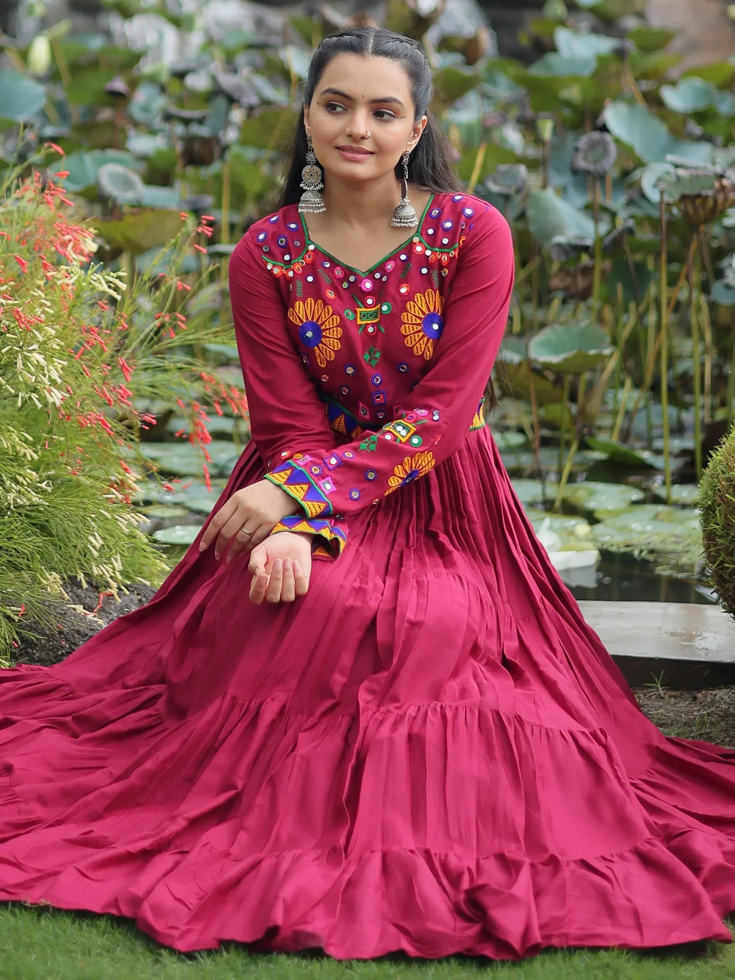 Maroon ethnic gown with mirror embroidery, floral motifs, and tiered skirt, styled with jhumka earrings in garden setting