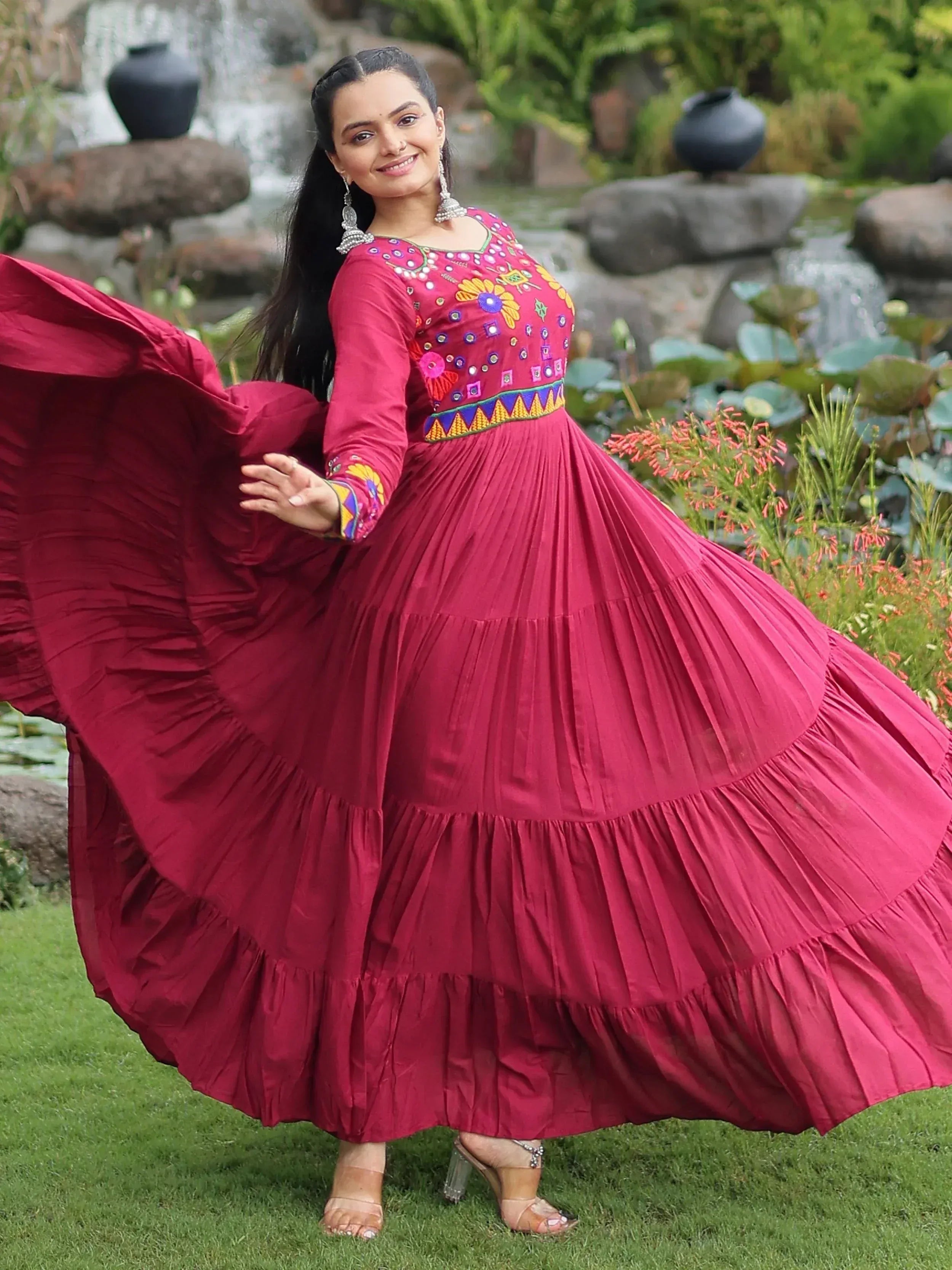 Woman in vibrant maroon ethnic gown with floral embroidery, twirling in garden setting