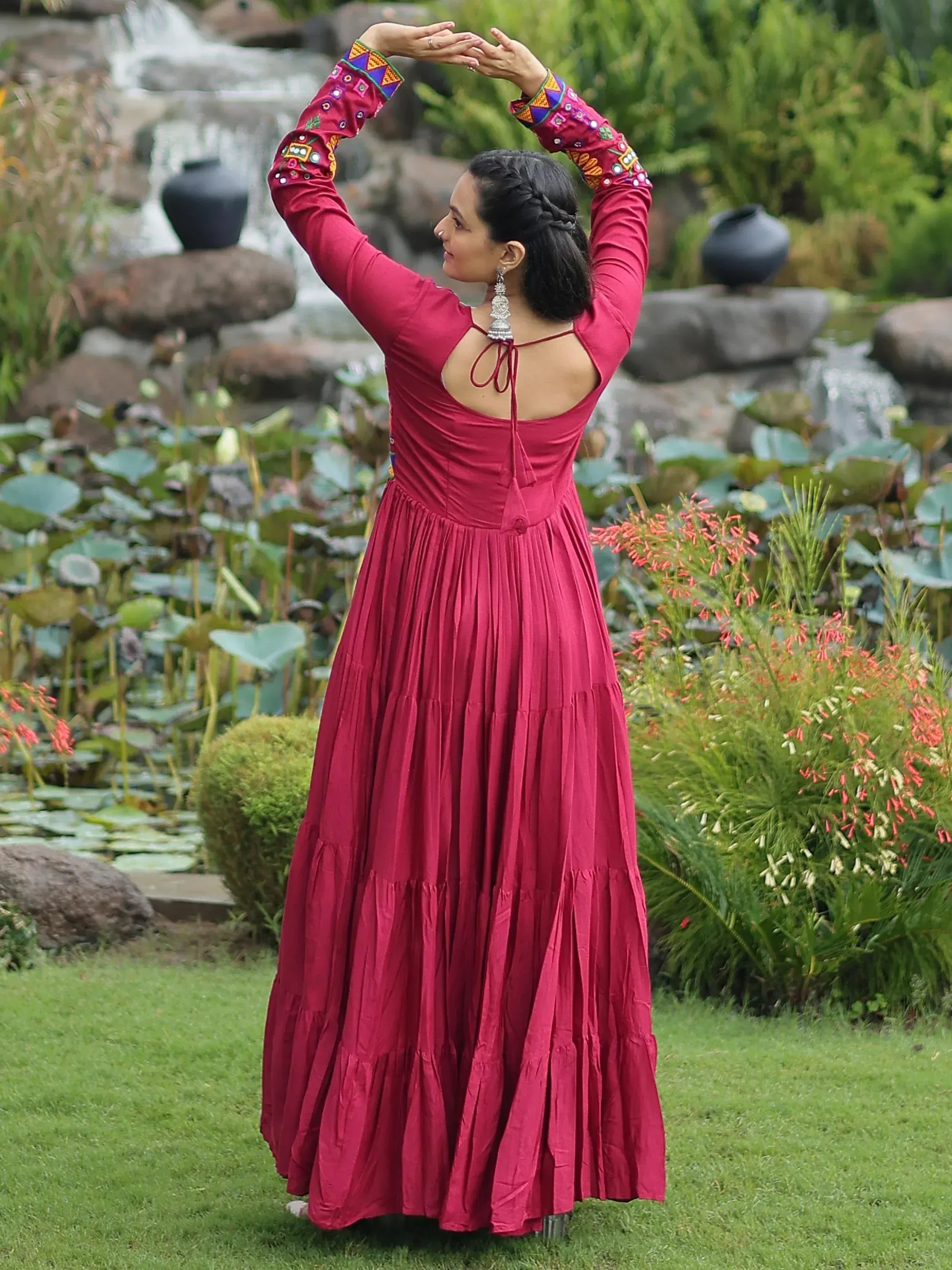 Woman in maroon ethnic gown with embroidered sleeves, standing in garden near pond