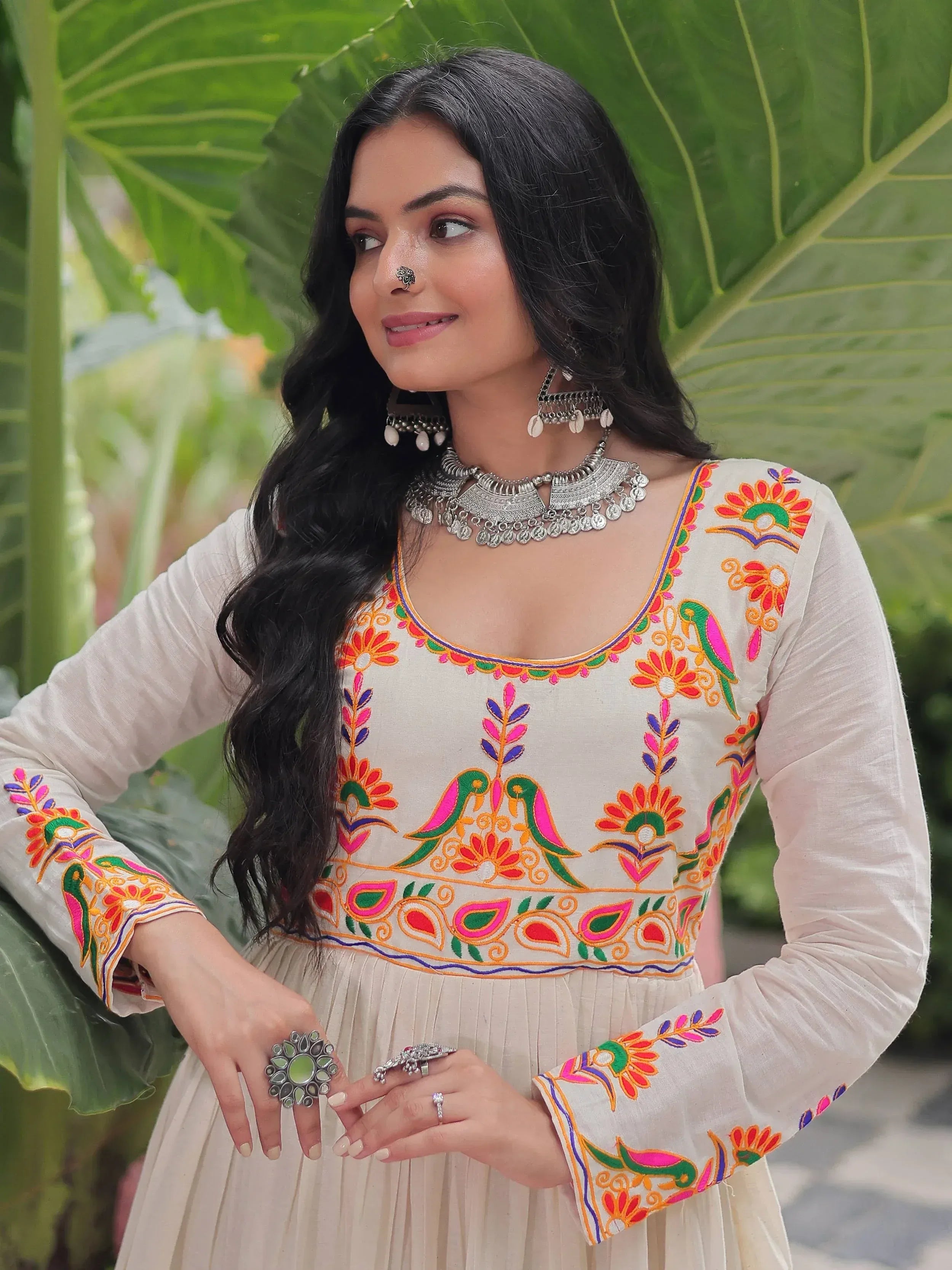 Woman in embroidered ethnic gown with colorful floral and bird motifs, silver jewelry, against green leaf backdrop