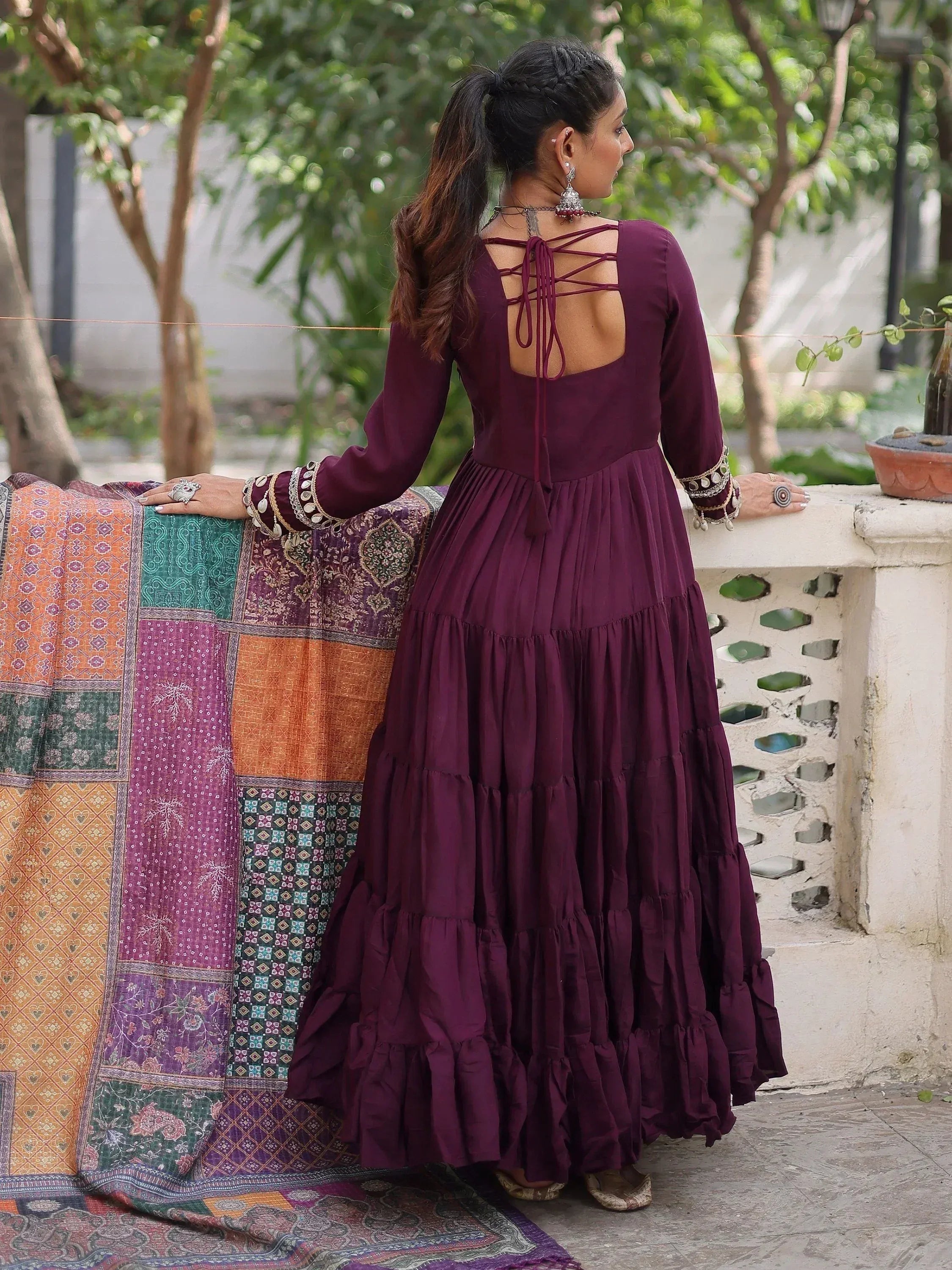 Purple ethnic gown with crisscross back, tiered skirt, and embellished sleeves, styled with traditional jewelry.