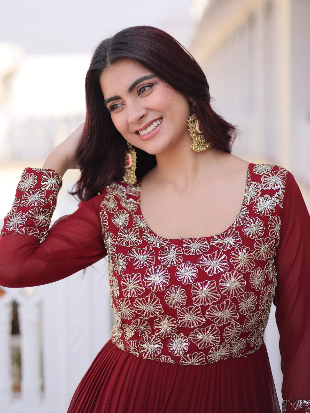 Woman wearing a maroon ethnic gown with gold floral embroidery and traditional jhumka earrings.