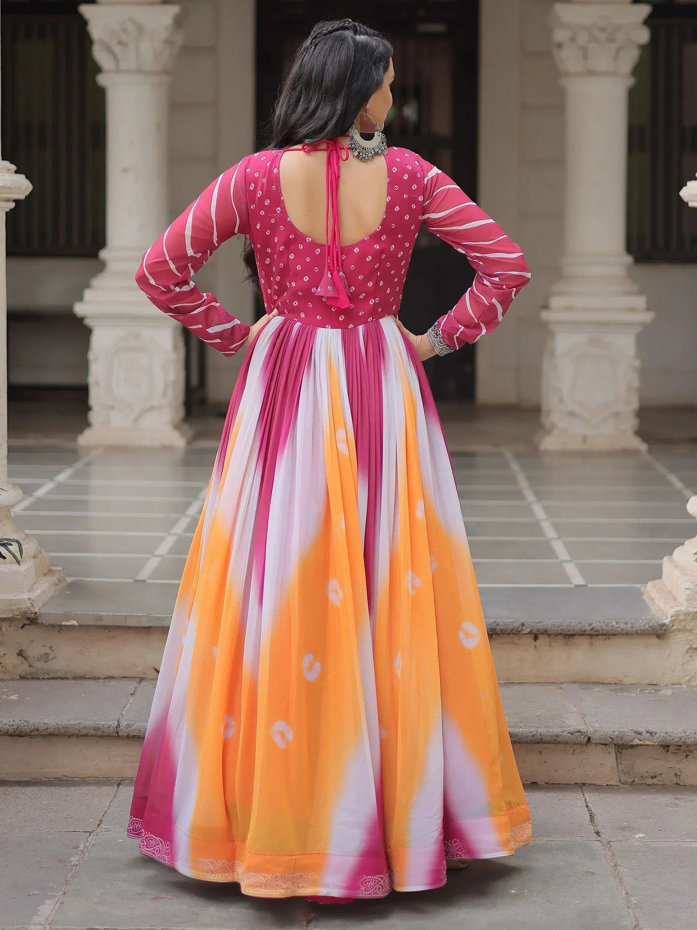 Woman in vibrant pink and orange tie-dye ethnic gown with back tie detail, standing near ornate pillars