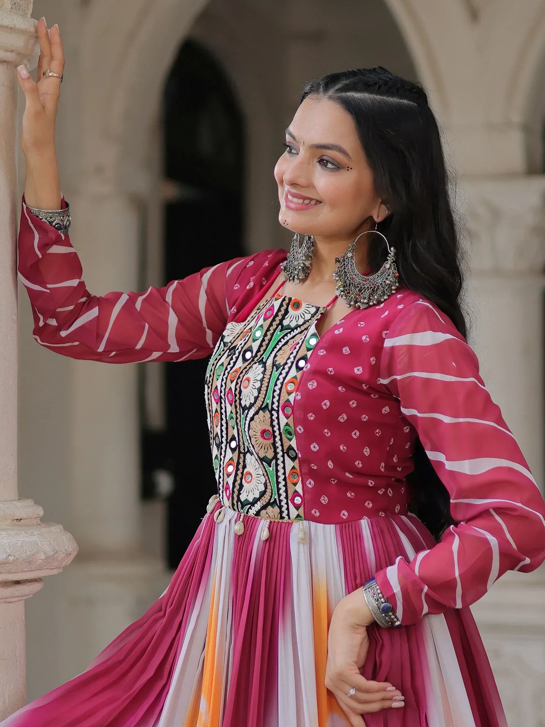 Woman in vibrant pink ethnic gown with mirror work, striped dupatta, and traditional jewelry, perfect for festive occasions.