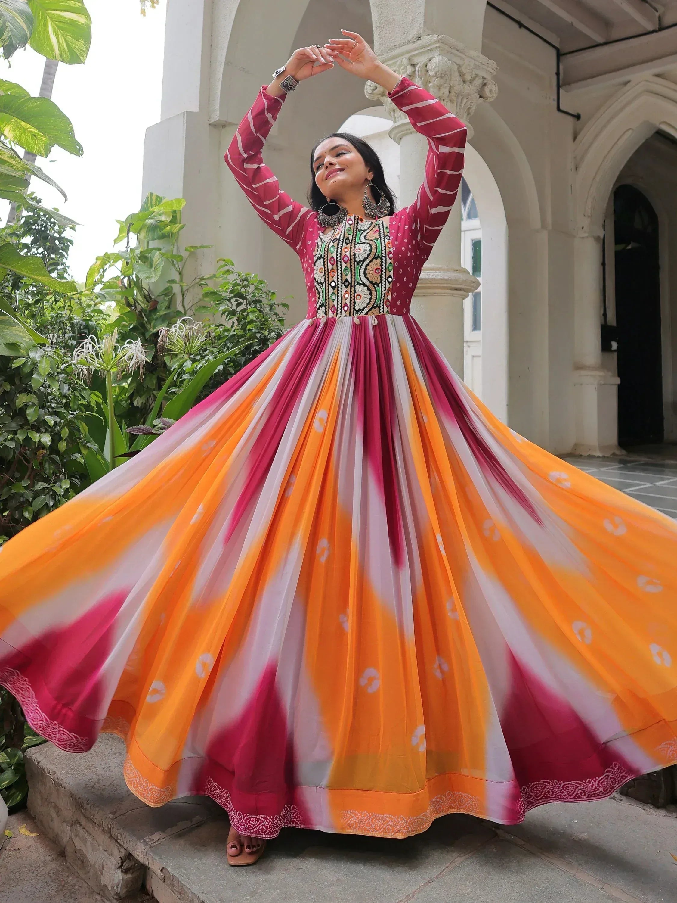 Woman in vibrant pink and orange ethnic gown with embroidered bodice, twirling outdoors near arches