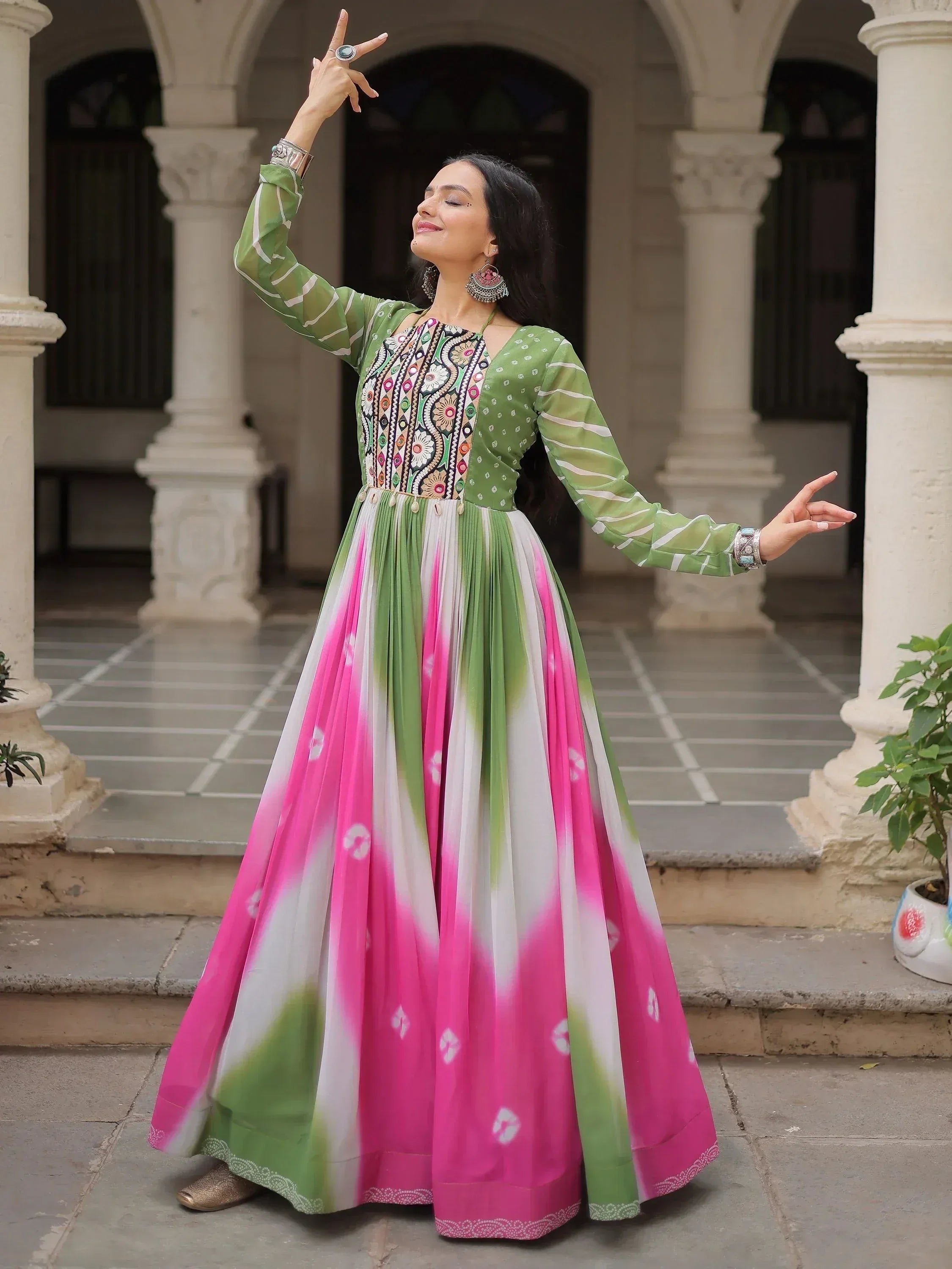 Woman in vibrant green and pink ethnic gown with floral embroidery, posing gracefully in courtyard.