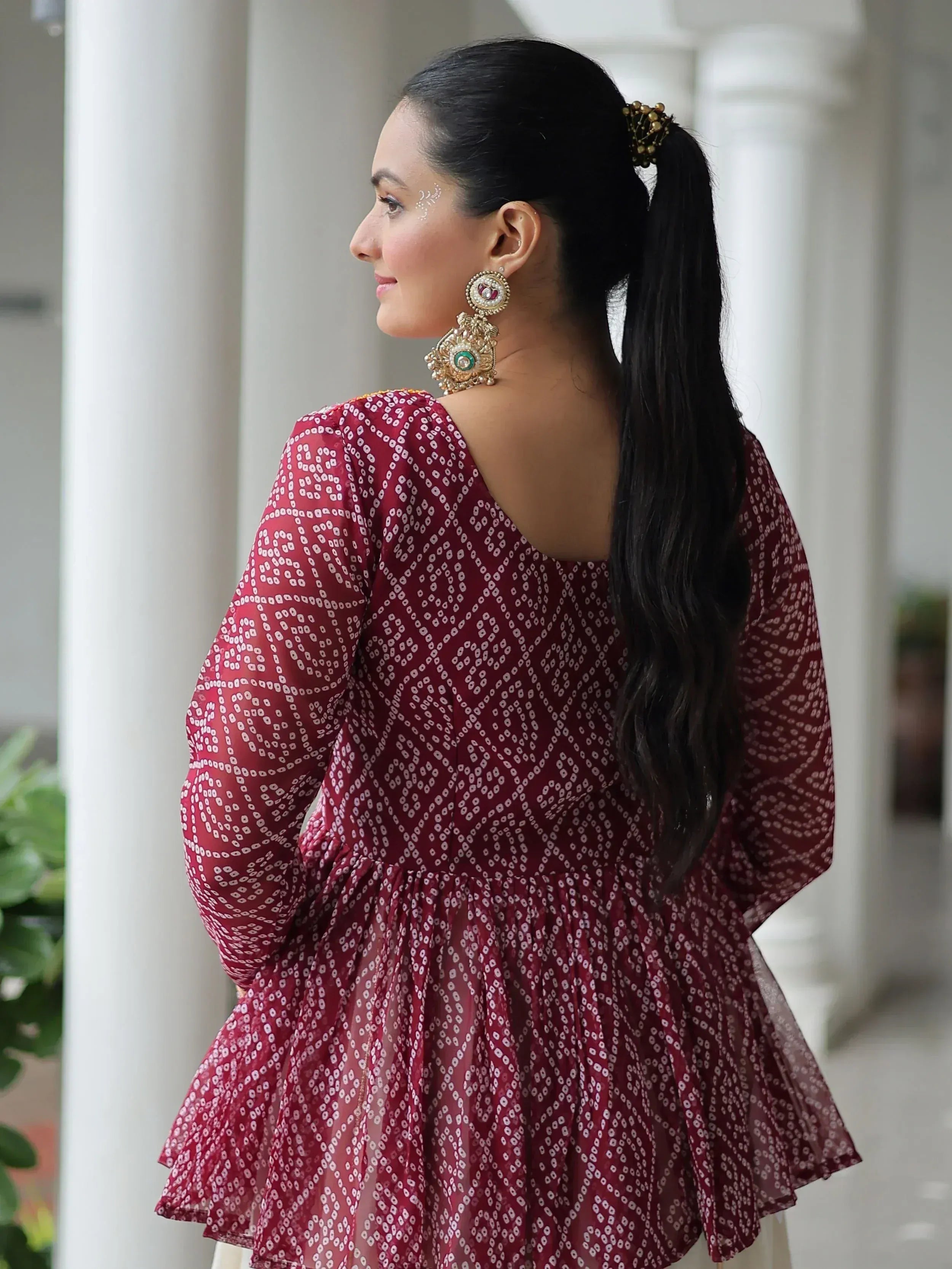 Woman in maroon ethnic gown with white print, statement earrings, and ponytail, showcasing Indian fashion.