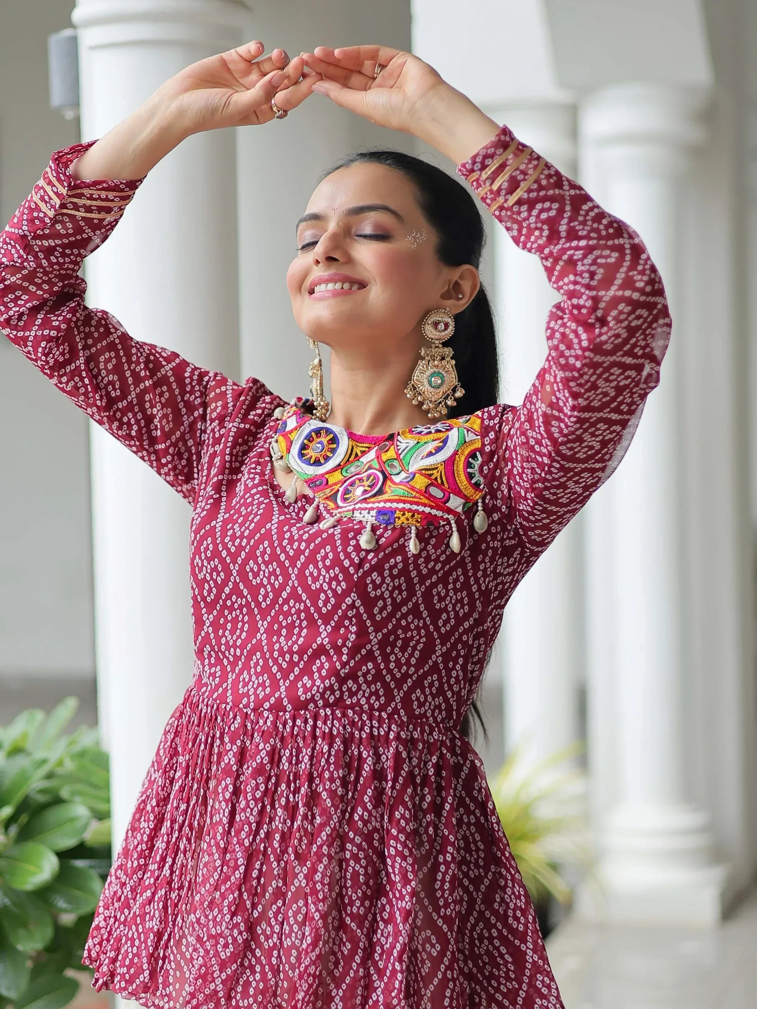 Woman in maroon ethnic dress with colorful embroidery and traditional jewelry, posing gracefully.