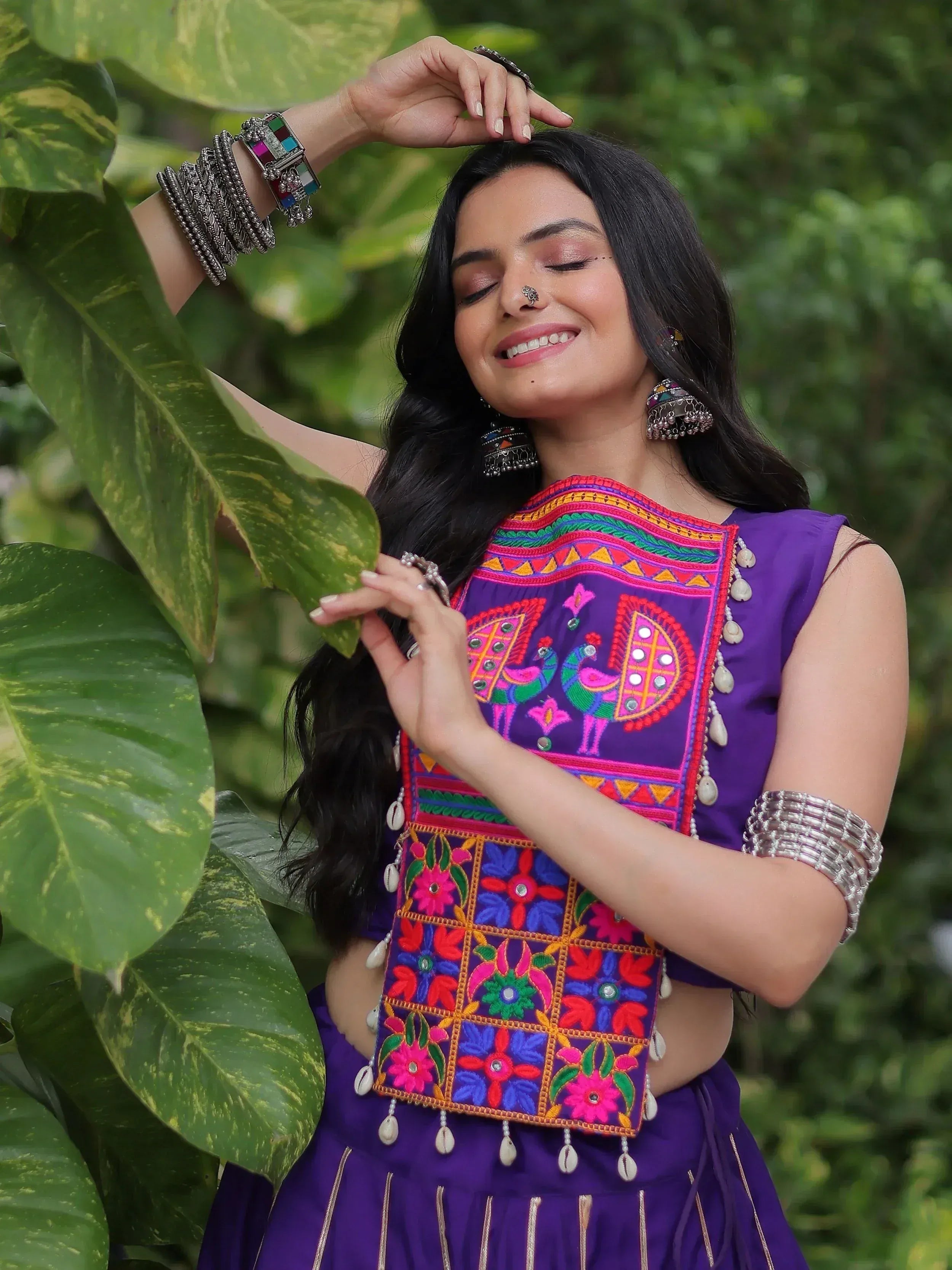 Woman in vibrant purple ethnic top with colorful peacock embroidery, floral patterns, and shell tassels, surrounded by green foliage.