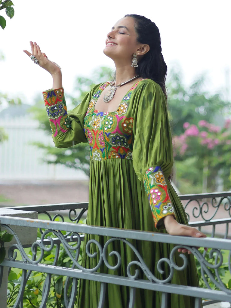 Woman in green embroidered ethnic gown on balcony, enjoying rain, with floral jewelry and traditional Indian attire.
