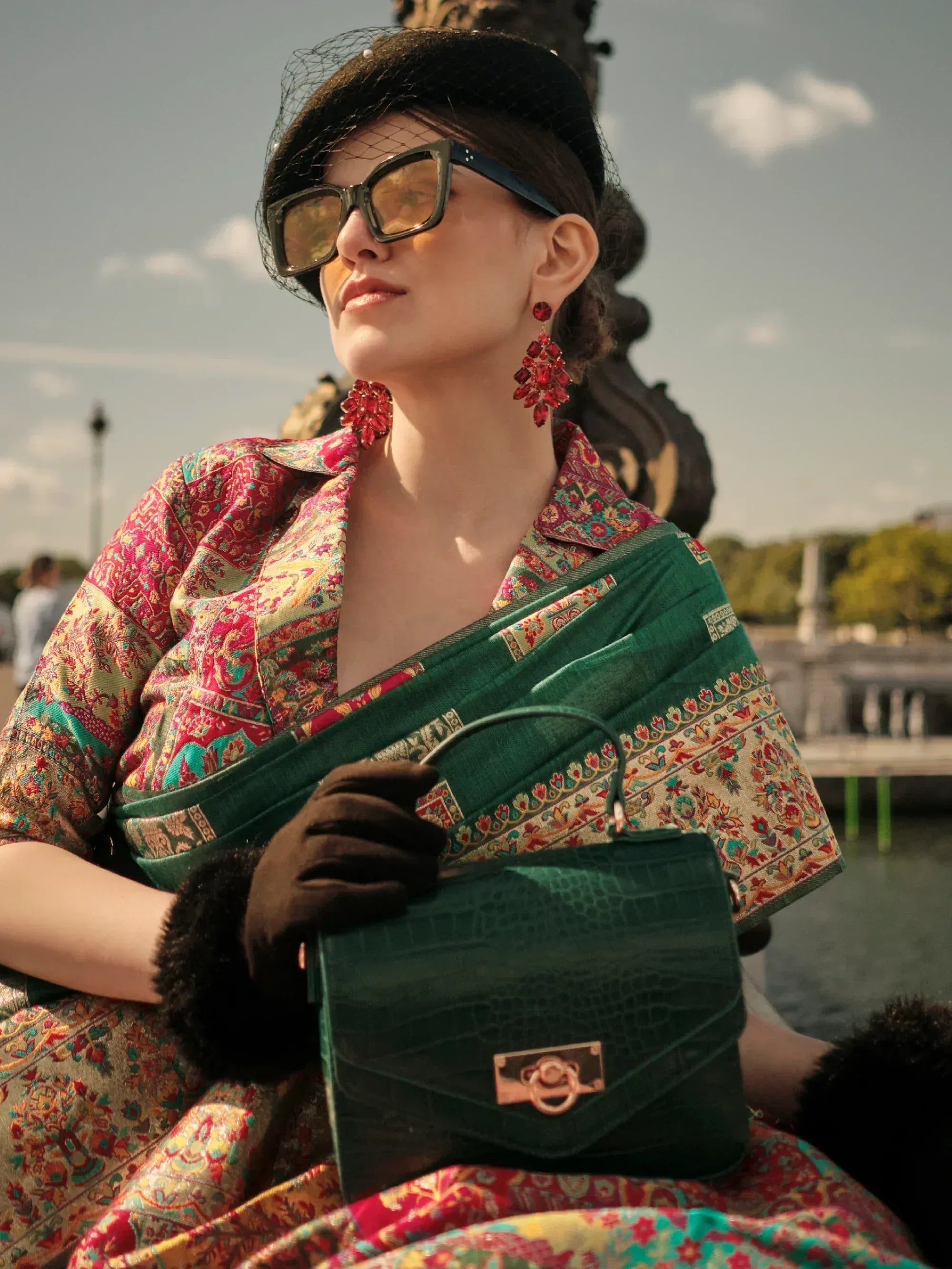 Woman in colorful Indian saree with green border, black hat, sunglasses, and statement earrings holding green handbag