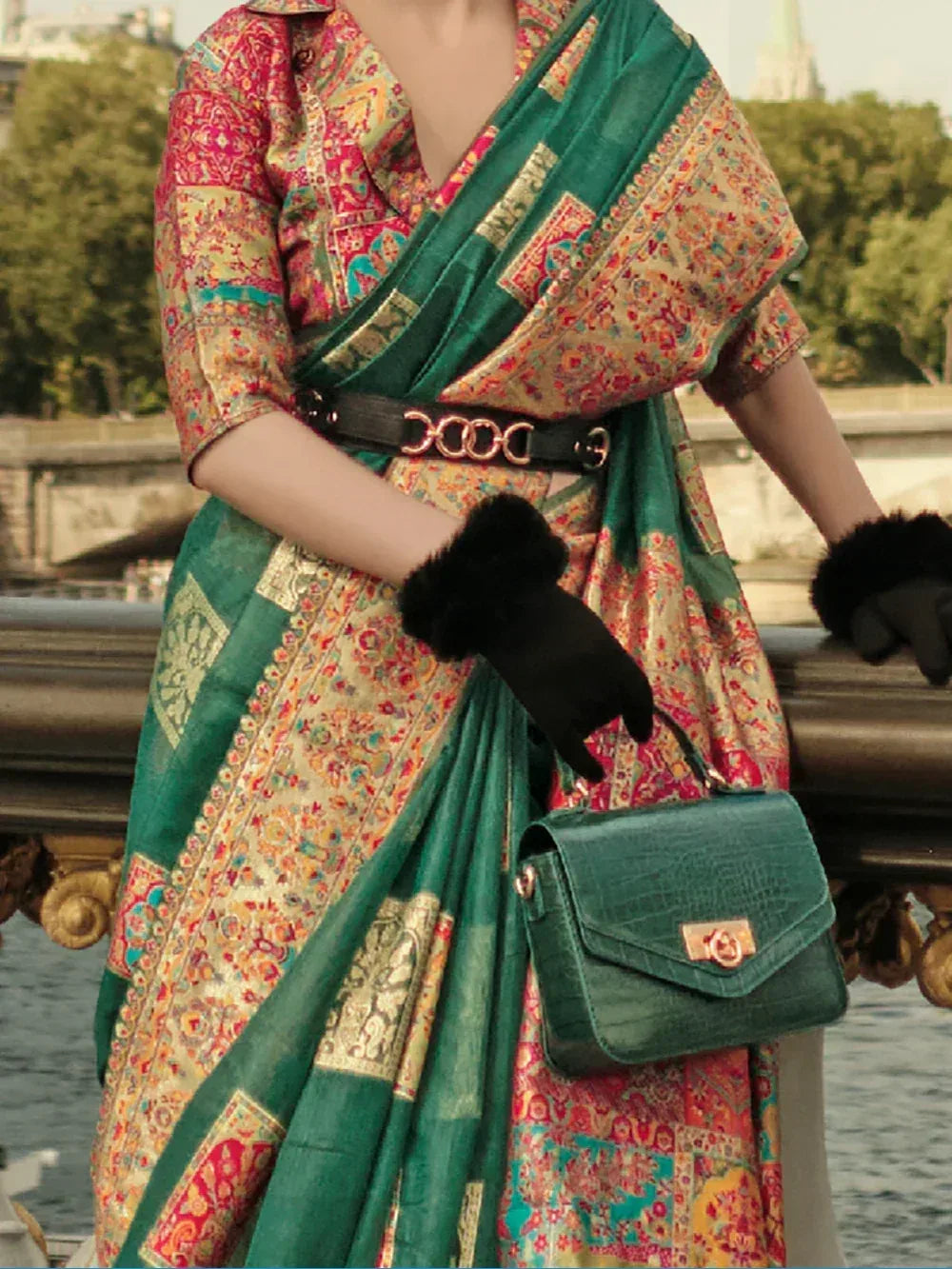 Woman in vibrant printed saree with green border, black belt, gloves, and matching handbag, styled for ethnic fashion.