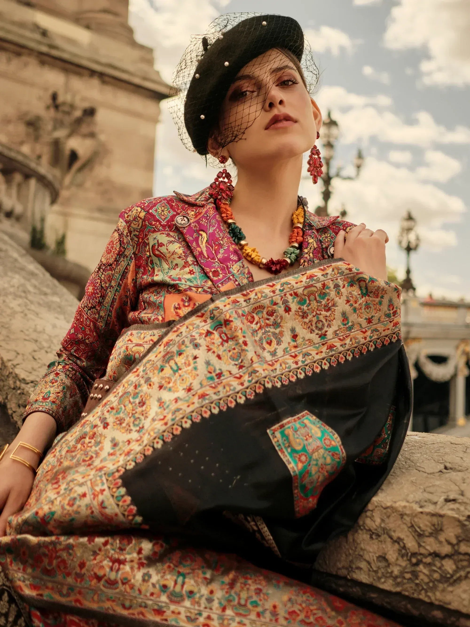 Woman in vibrant ethnic print salwar kameez with black beret, statement jewelry, and ornate dupatta, styled for festive occasions.