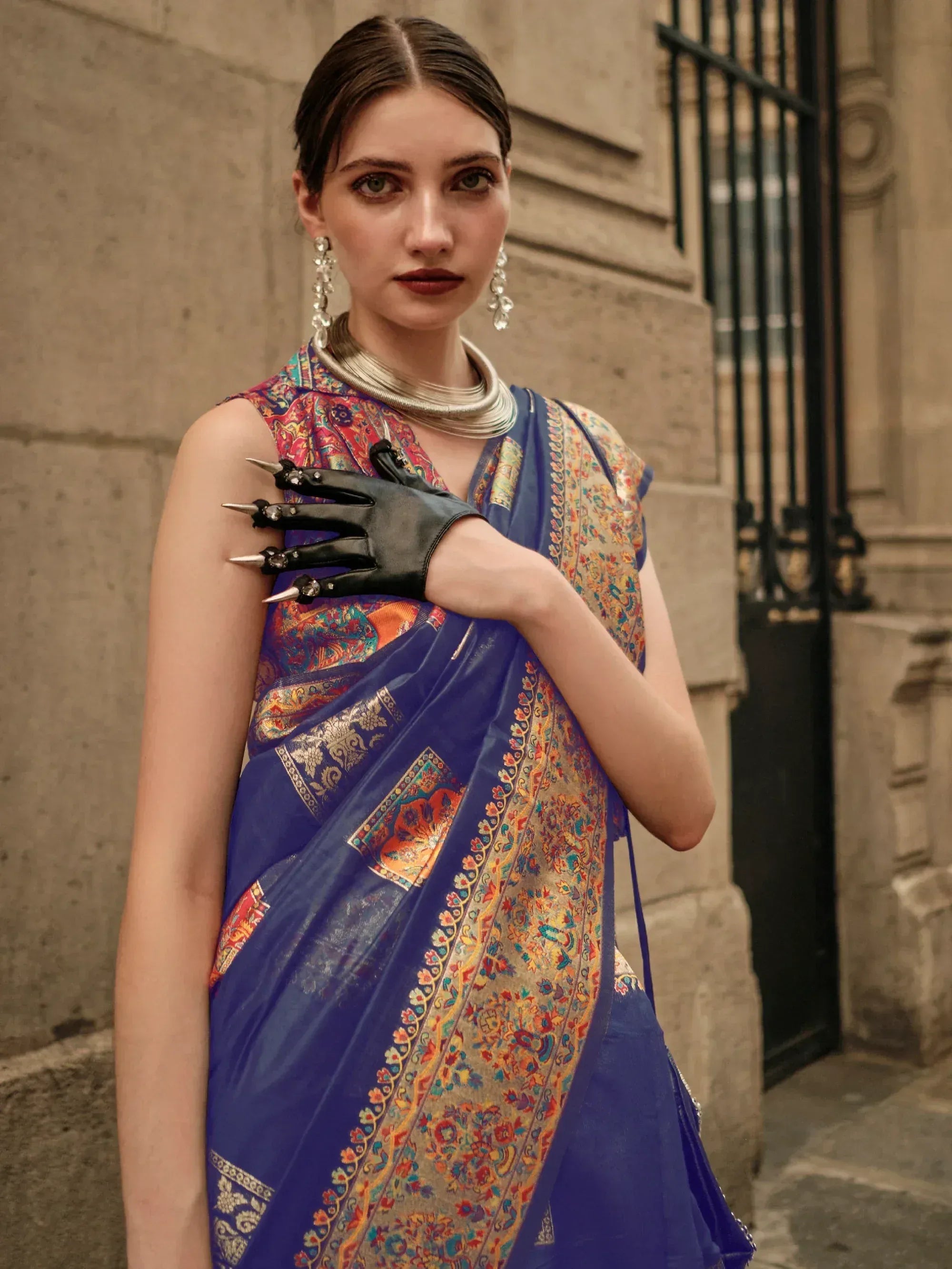 Woman in blue printed saree with gold border, statement jewelry, and black spiked gloves, styled for ethnic fashion.