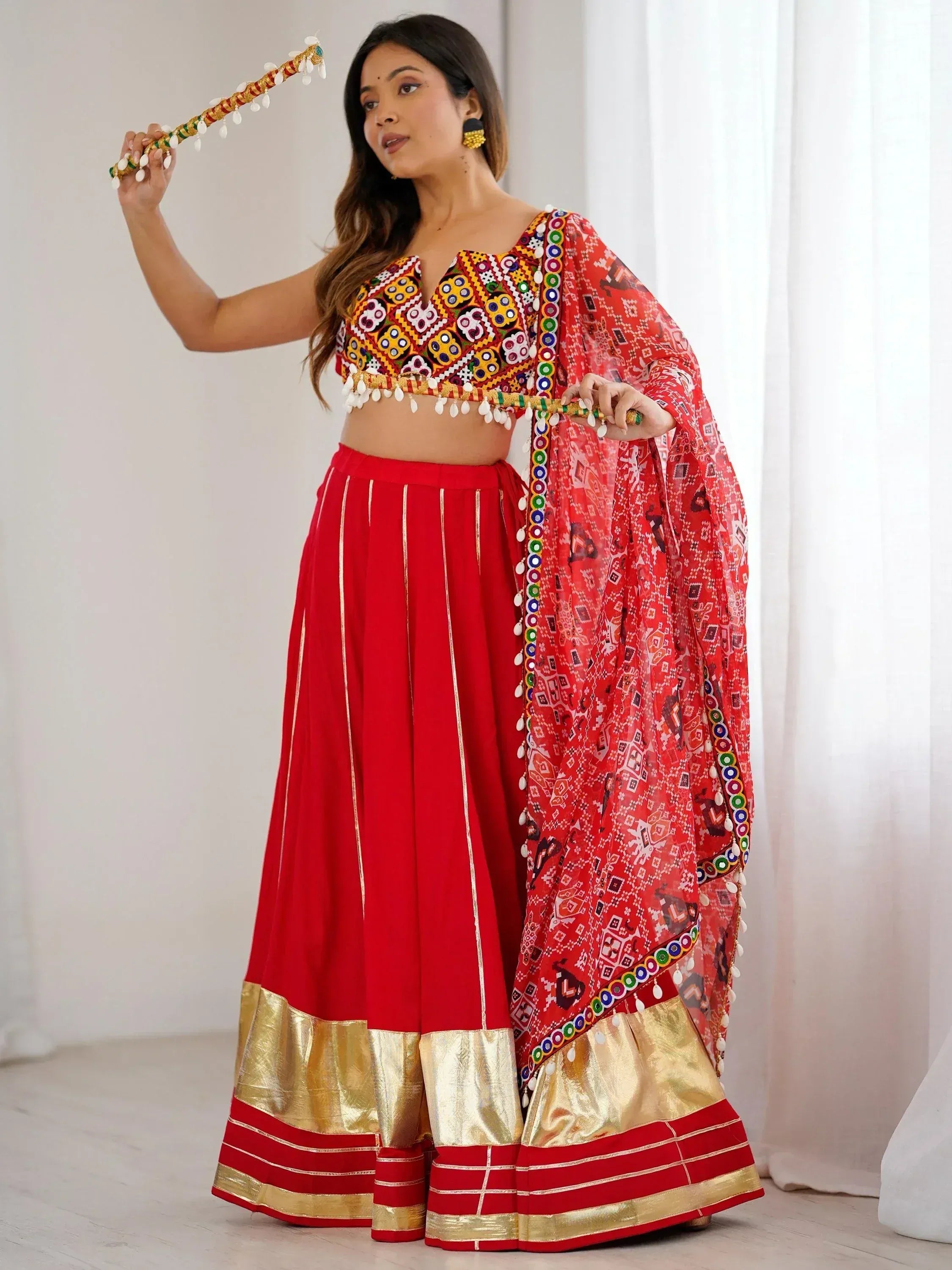 Red embroidered lehenga choli with mirror work, gold border, and dupatta, styled with traditional dandiya stick for festive occasions