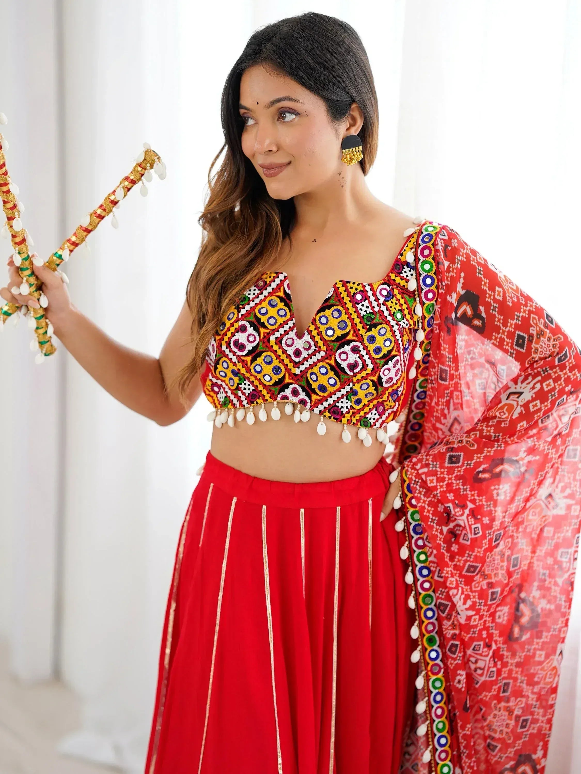 Woman in vibrant red ethnic lehenga choli with mirror work, holding decorative dandiya sticks for festive occasions.
