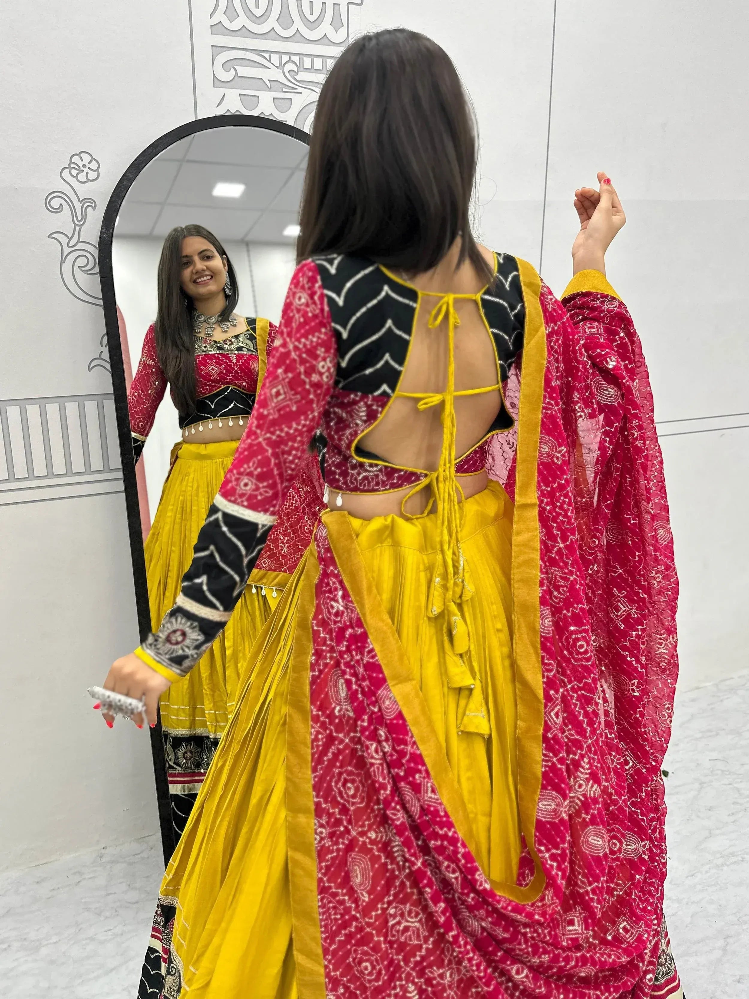 Woman in vibrant red and yellow ethnic lehenga choli with back tie detail, checking mirror reflection