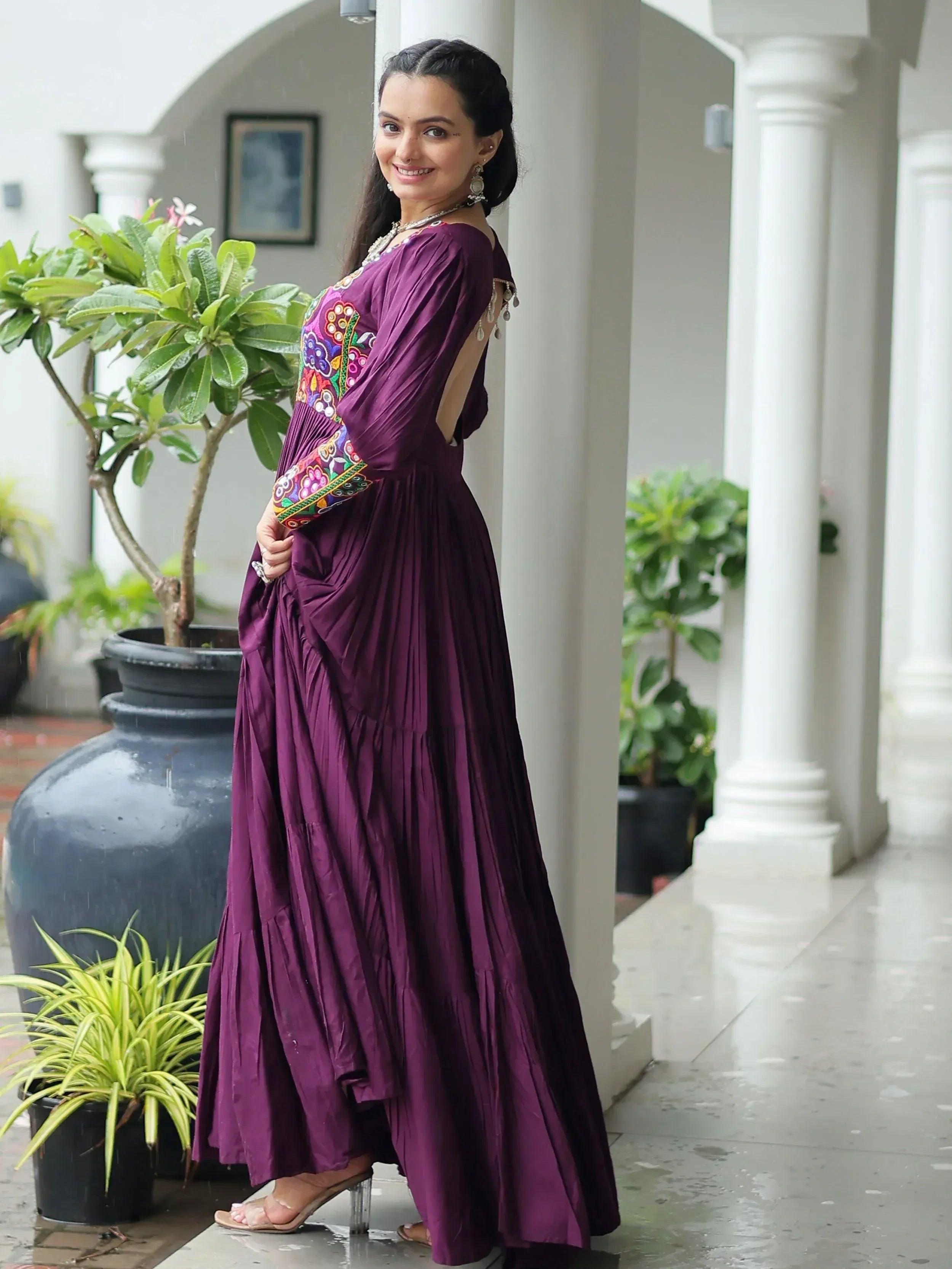 Purple ethnic gown with colorful floral embroidery, pleated skirt, and back cutout, styled with statement jewelry.