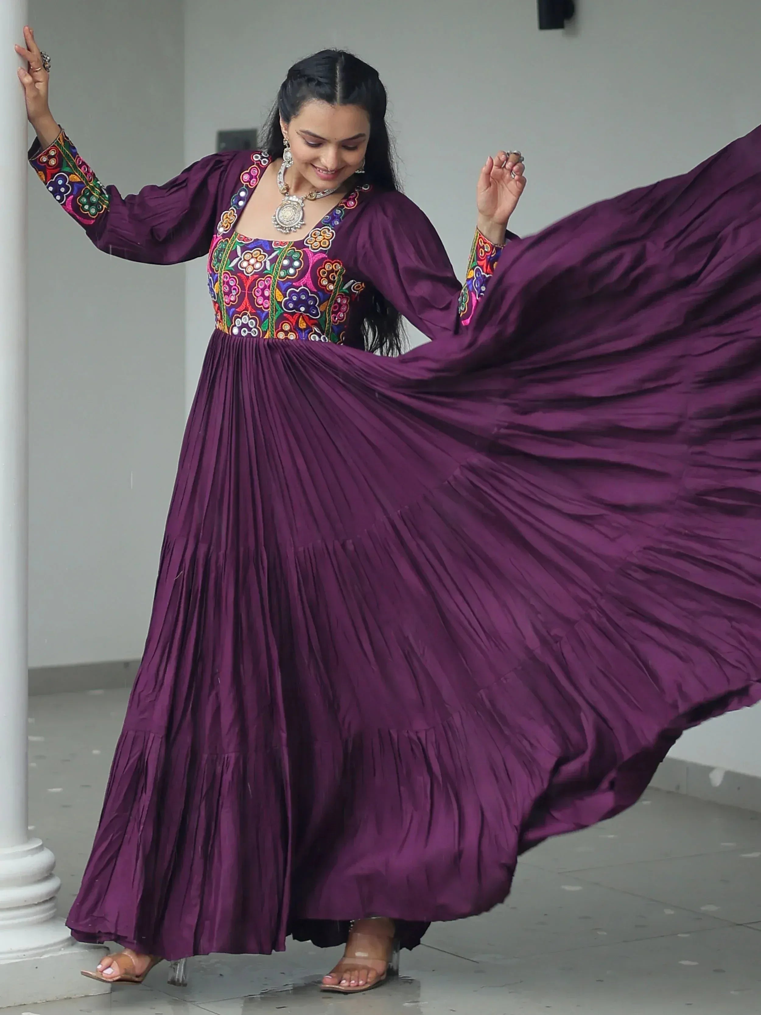 Woman in purple ethnic gown with colorful floral embroidery, pleated skirt, and silver jewelry.