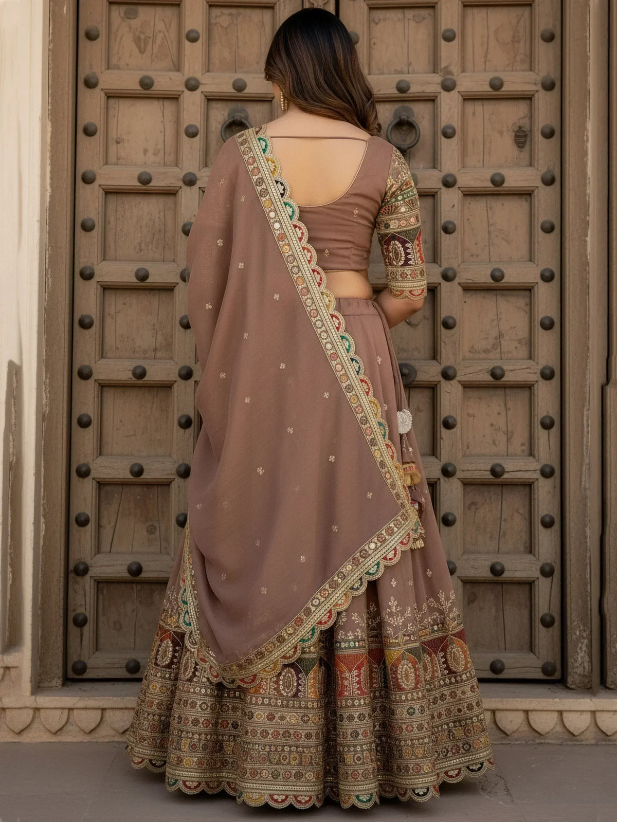 Back view of woman in designer lehenga choli with intricate embroidery, standing before rustic wooden door
