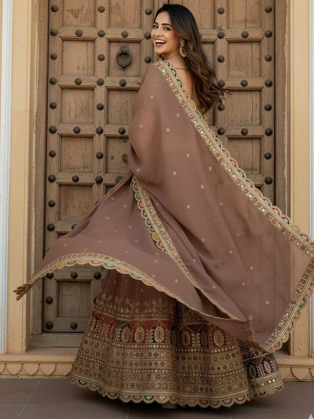 Woman in elegant brown embroidered lehenga choli with scalloped dupatta, standing before rustic wooden door