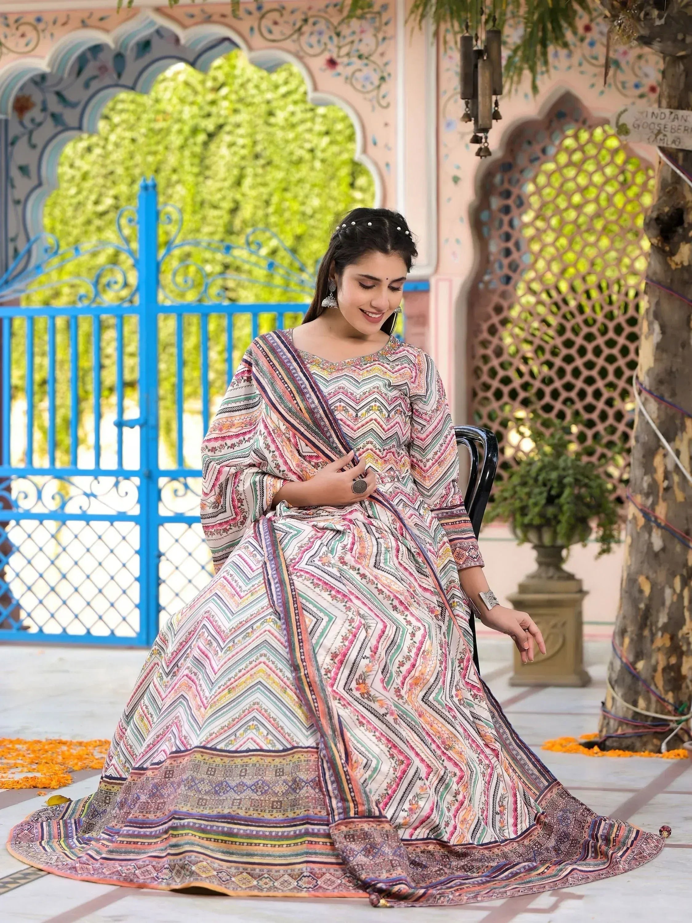 Woman in colorful Indian ethnic gown with geometric print, seated near ornate gate and pink wall