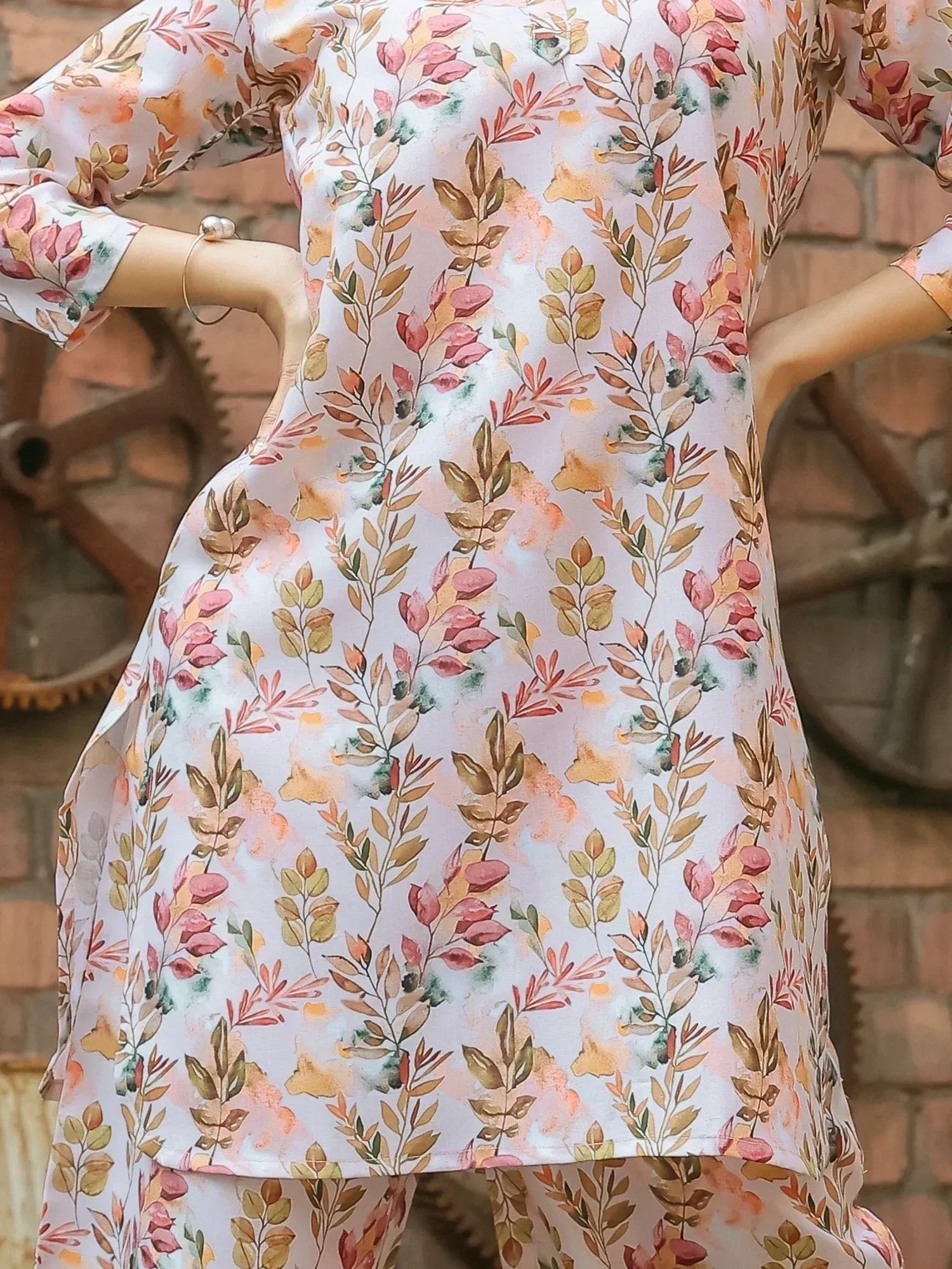 Woman wearing pastel floral print kurti and pants, standing near brick wall with gears