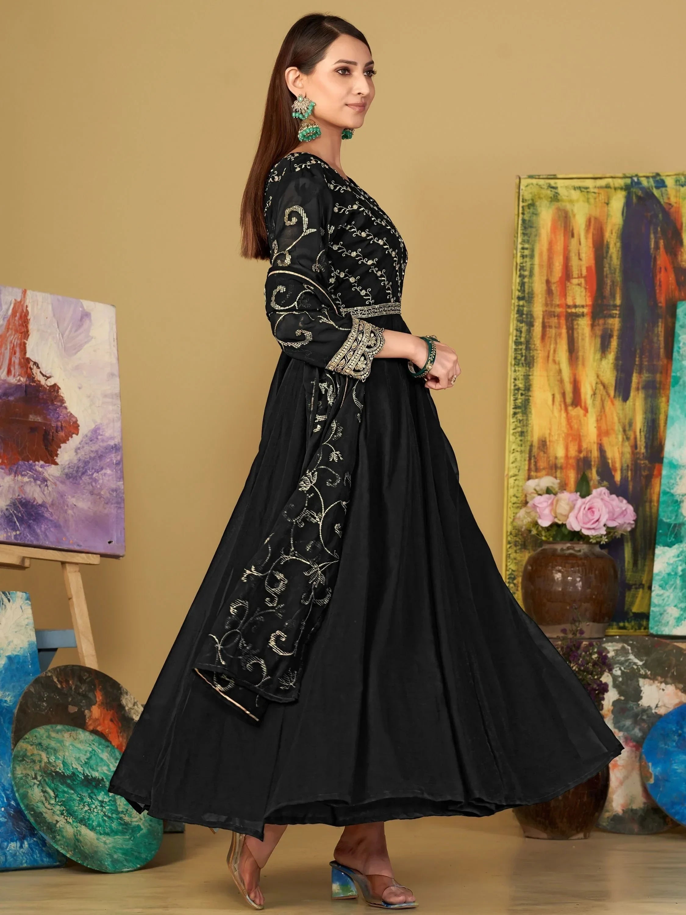 Woman in black embroidered ethnic gown with floral earrings, standing near colorful paintings