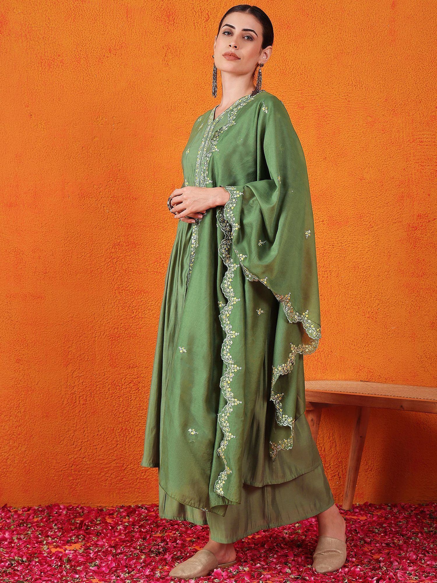 Woman wearing green embroidered ethnic gown with dupatta, standing against orange wall