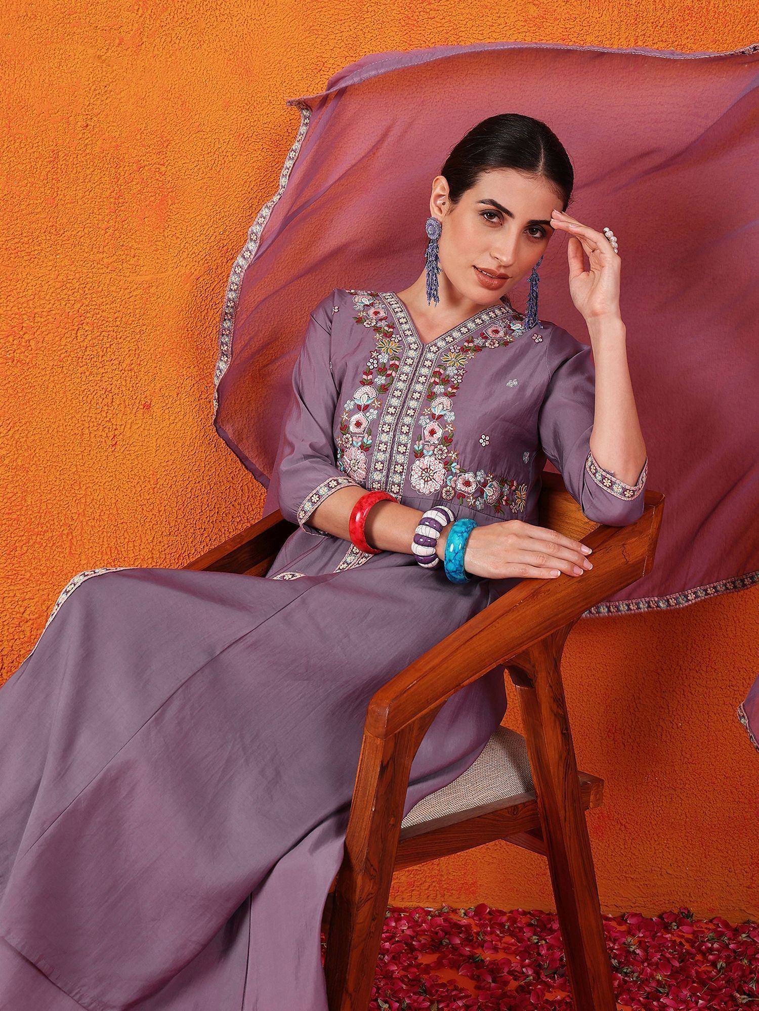 Woman in purple embroidered ethnic gown with floral details, styled with bangles and earrings, seated on wooden chair.