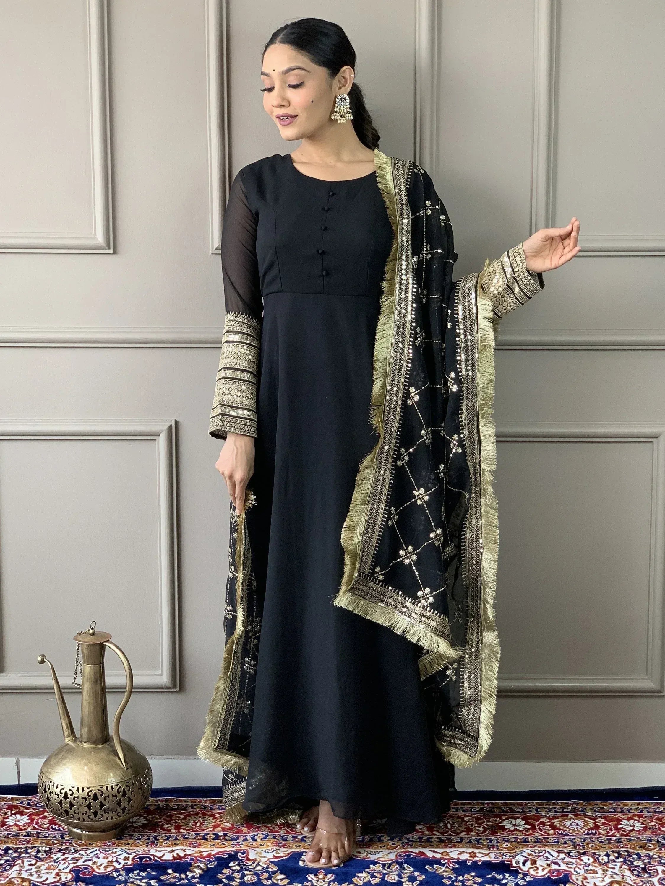 Black ethnic gown with gold embroidery and matching dupatta, styled with traditional earrings and a patterned rug
