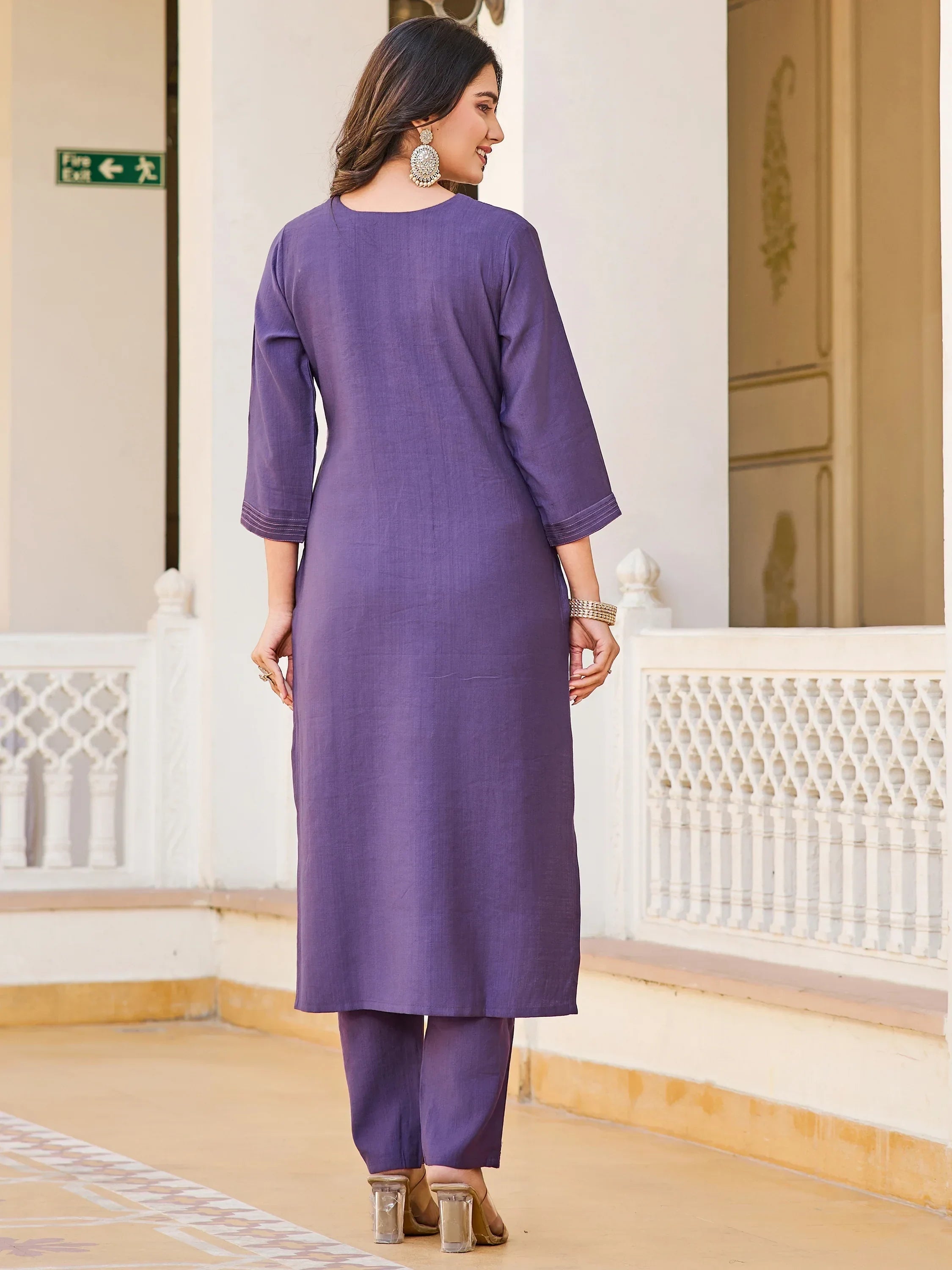 Woman in purple straight-cut kurti and pants, ethnic jewelry, standing in traditional setting